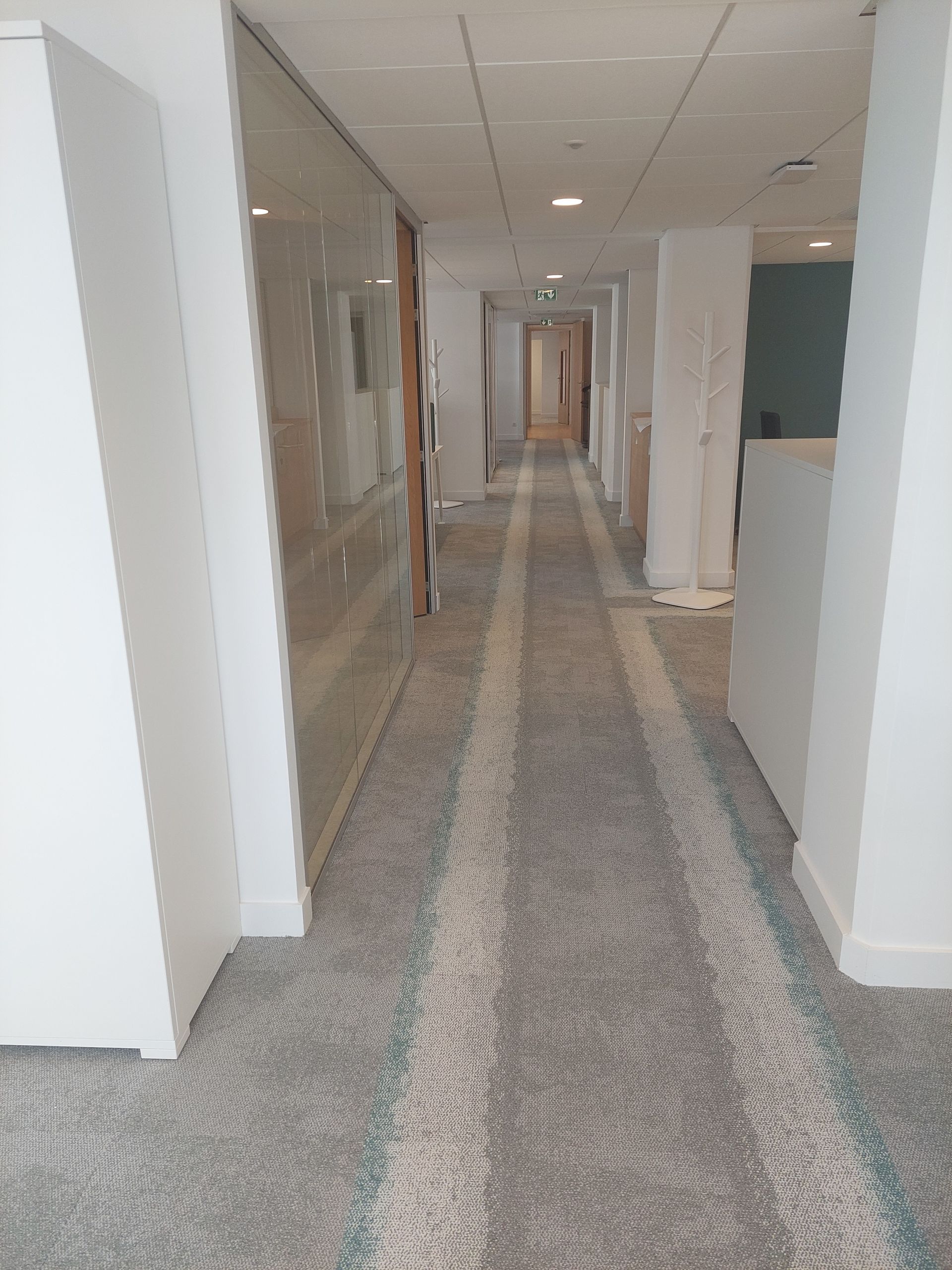 Long couloir aux murs blancs, au sol recouvert de moquette et avec éclairage encastré.