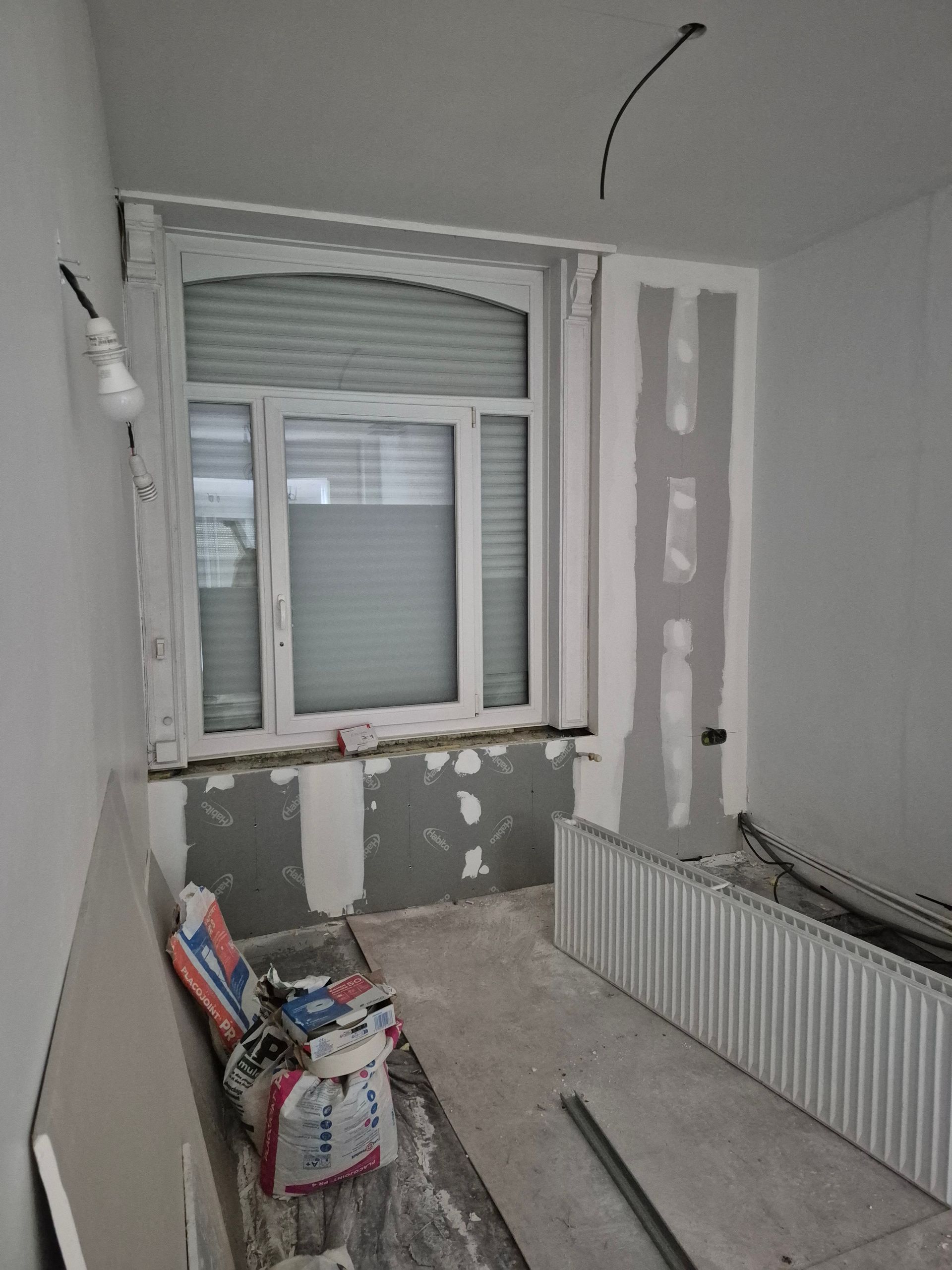 Pièce intérieure en rénovation, fenêtre avec stores, cloisons sèches apparentes, radiateur, matériaux de construction.