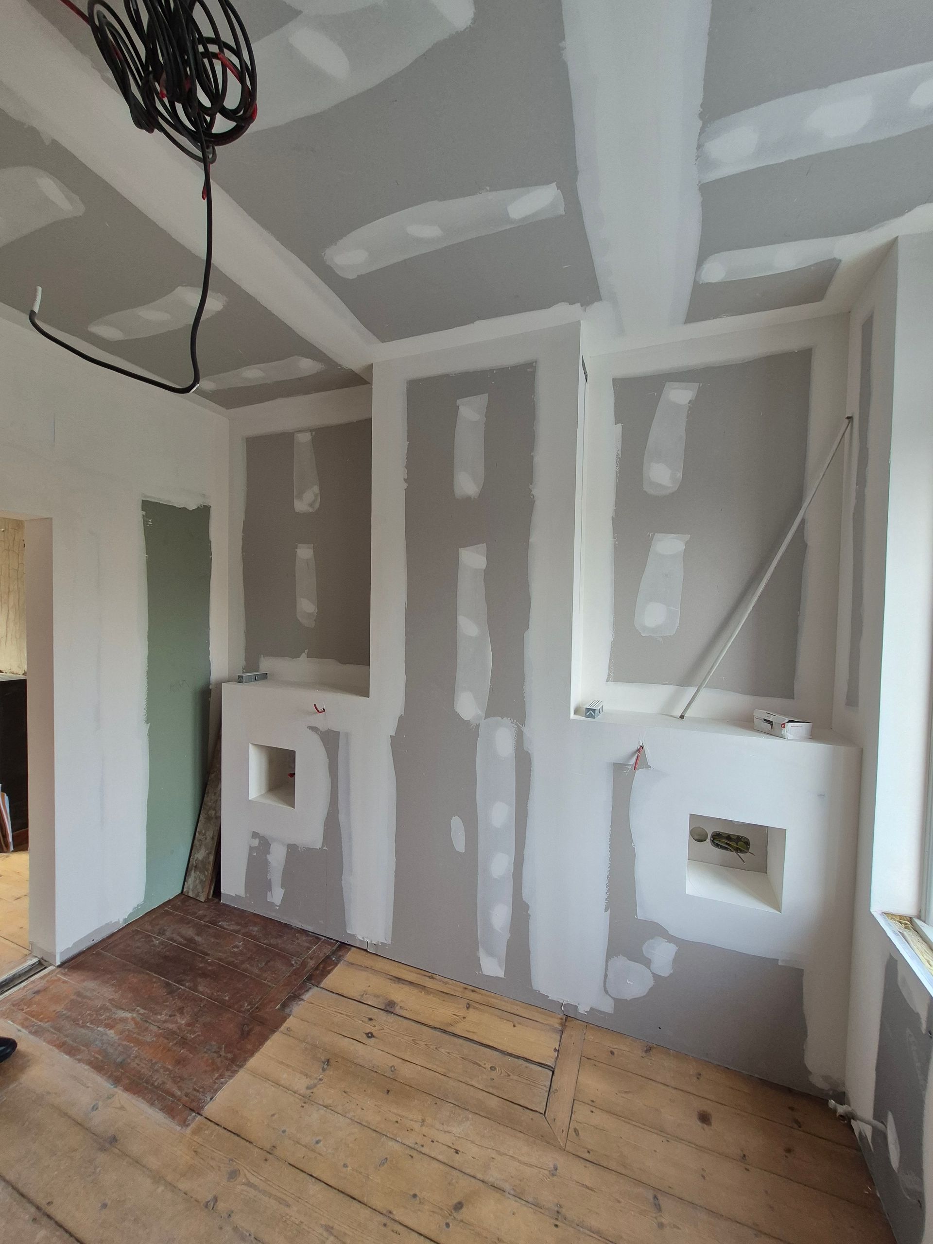 Pièce en travaux avec cloisons sèches apparentes et plancher en bois ; des fils électriques pendent du plafond.