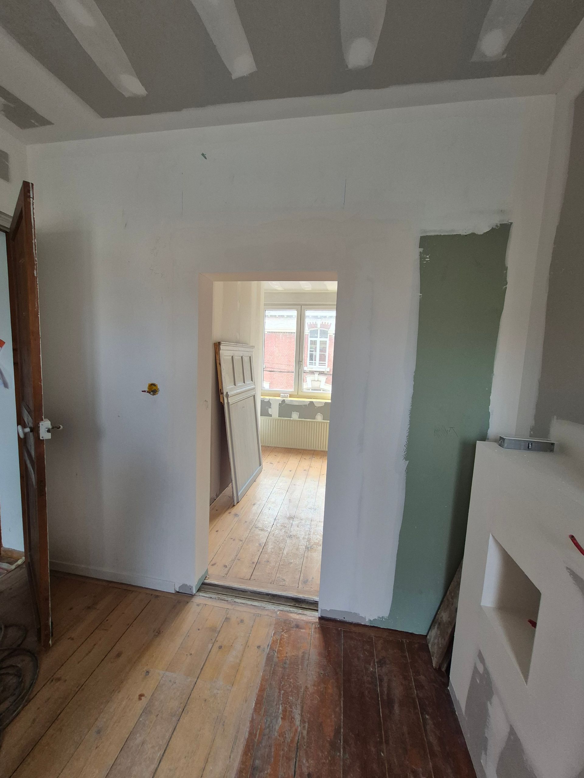 Intérieur en cours de rénovation ; une porte ouverte donne sur la cuisine avec fenêtre. Parquet, cloisons sèches et encadrement de porte apparents.