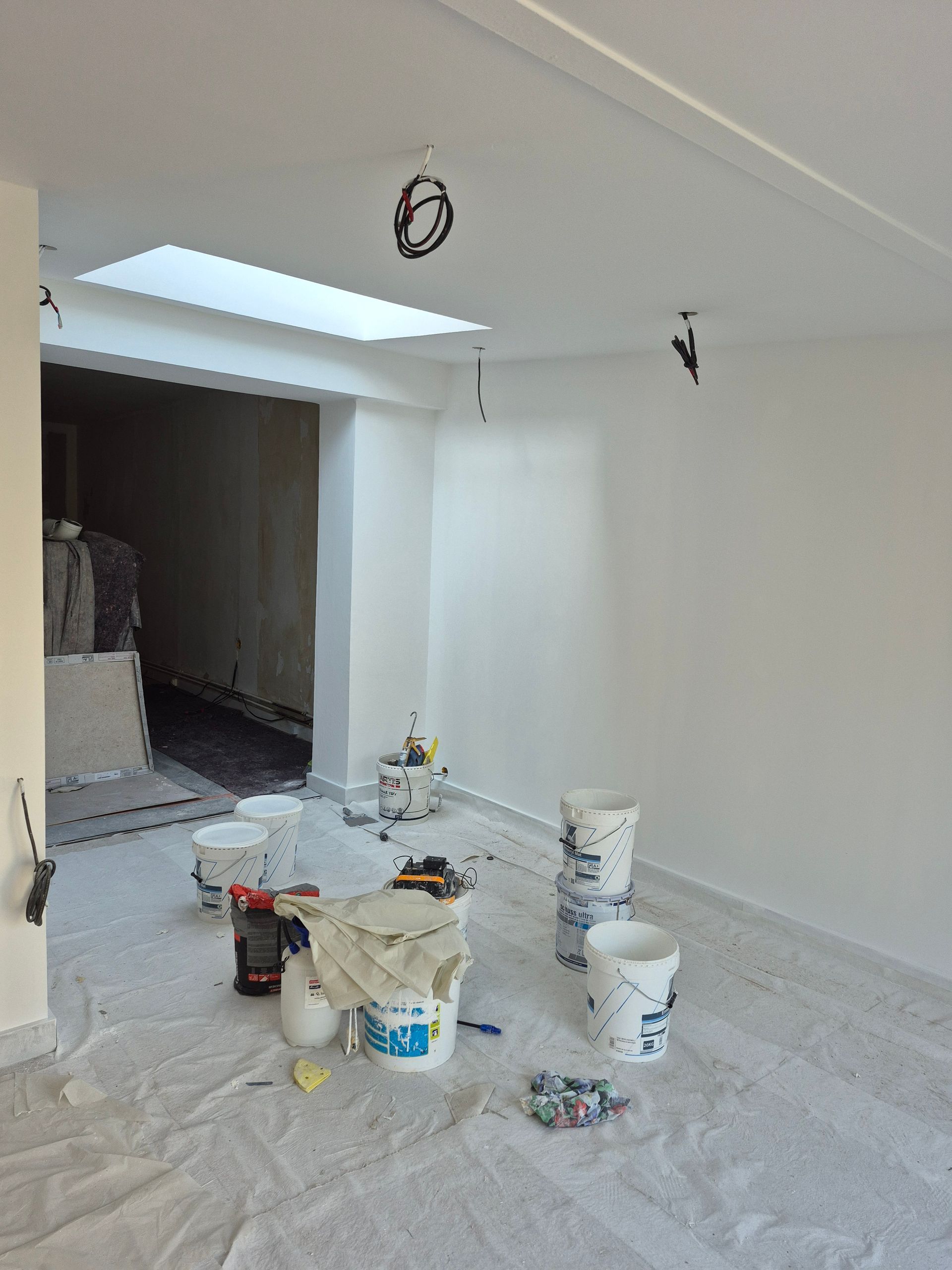 Pièce en rénovation ; murs blancs, seaux de peinture, bâche en plastique au sol. Puits de lumière au plafond.