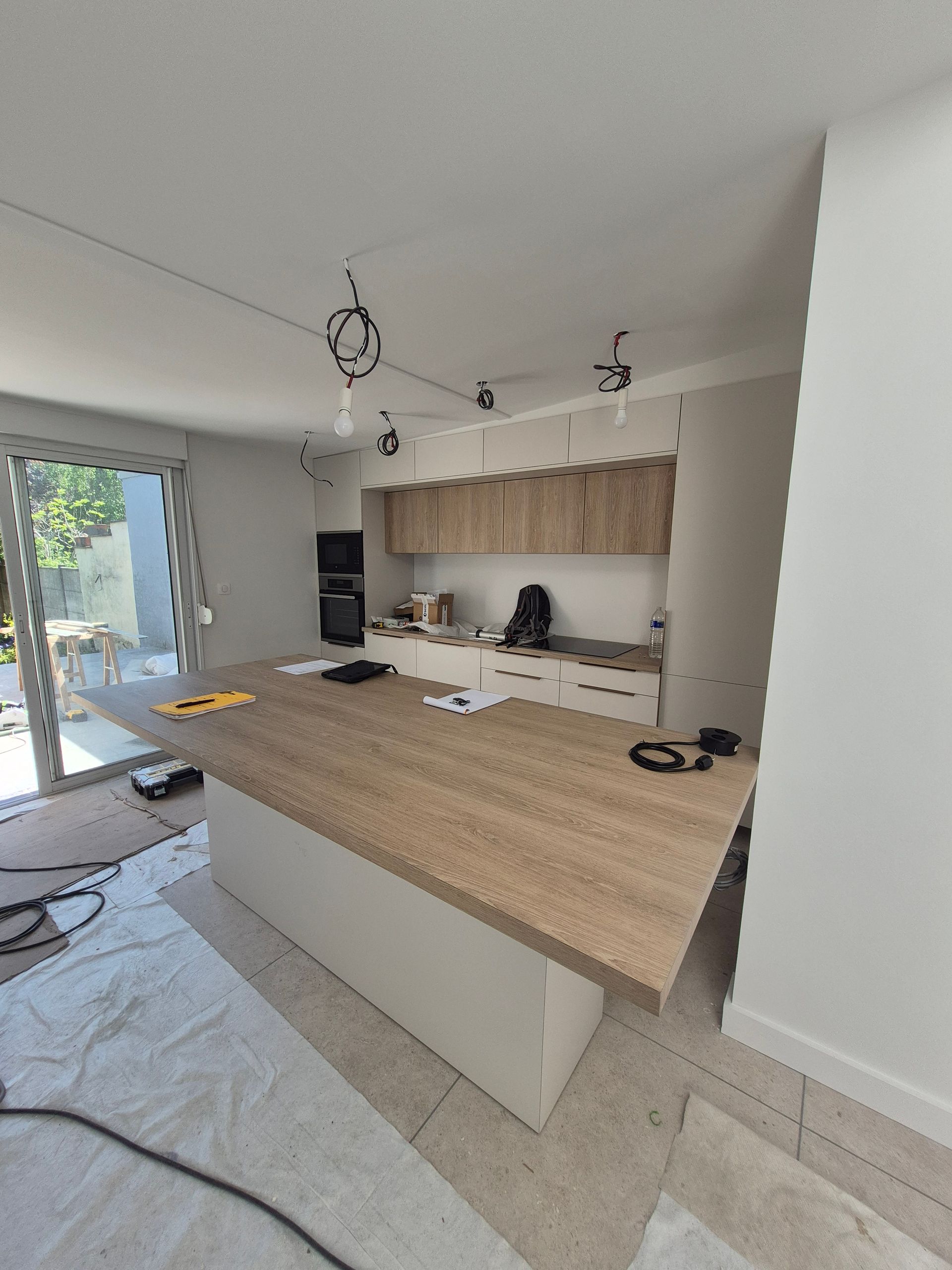 Rénovation de cuisine en cours : îlot avec comptoir en bois, armoires hautes, luminaires apparents, portes coulissantes en verre.