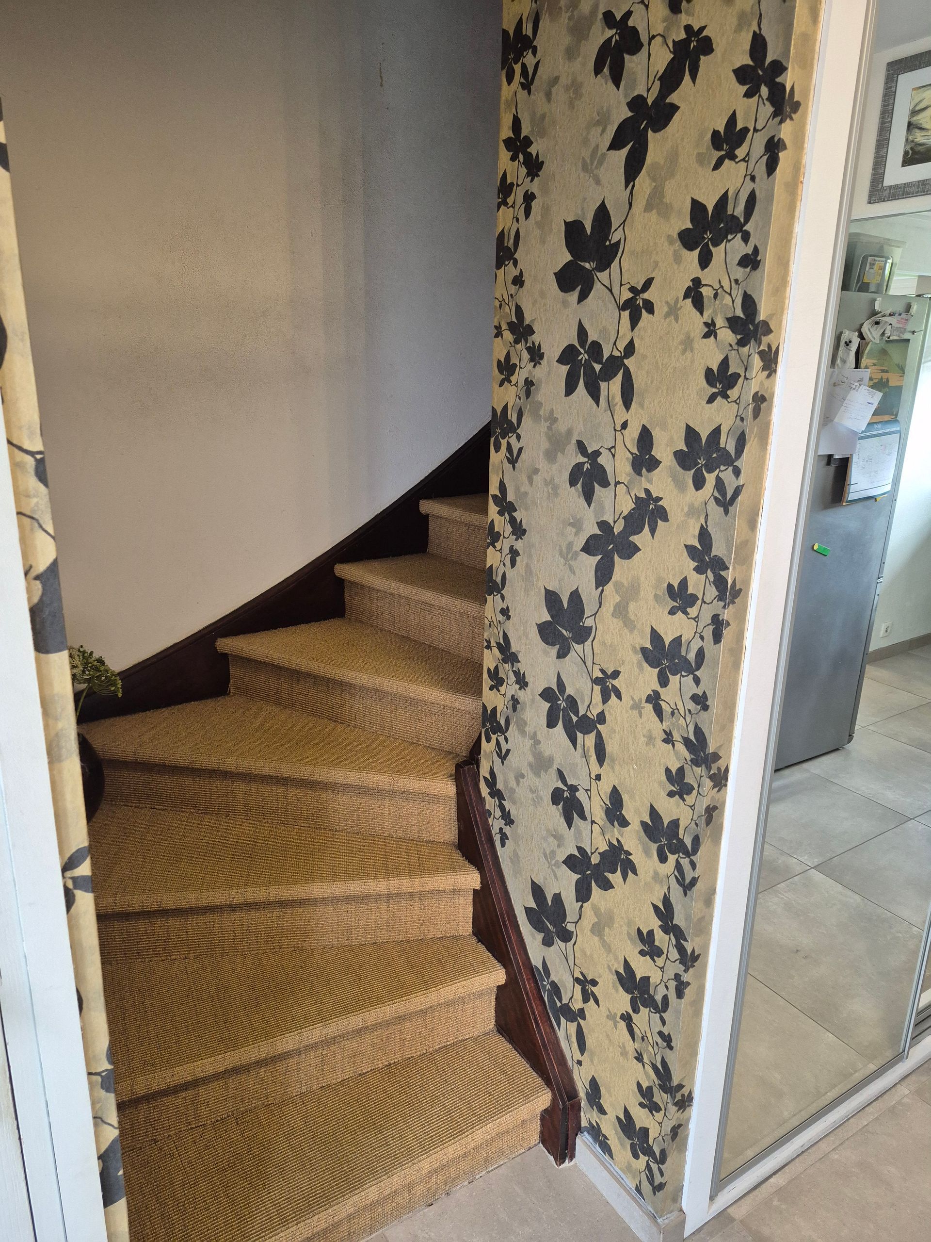 Escalier avec marches tressées, contremarches brun foncé et papier peint à motifs floraux, à côté d'un mur blanc.