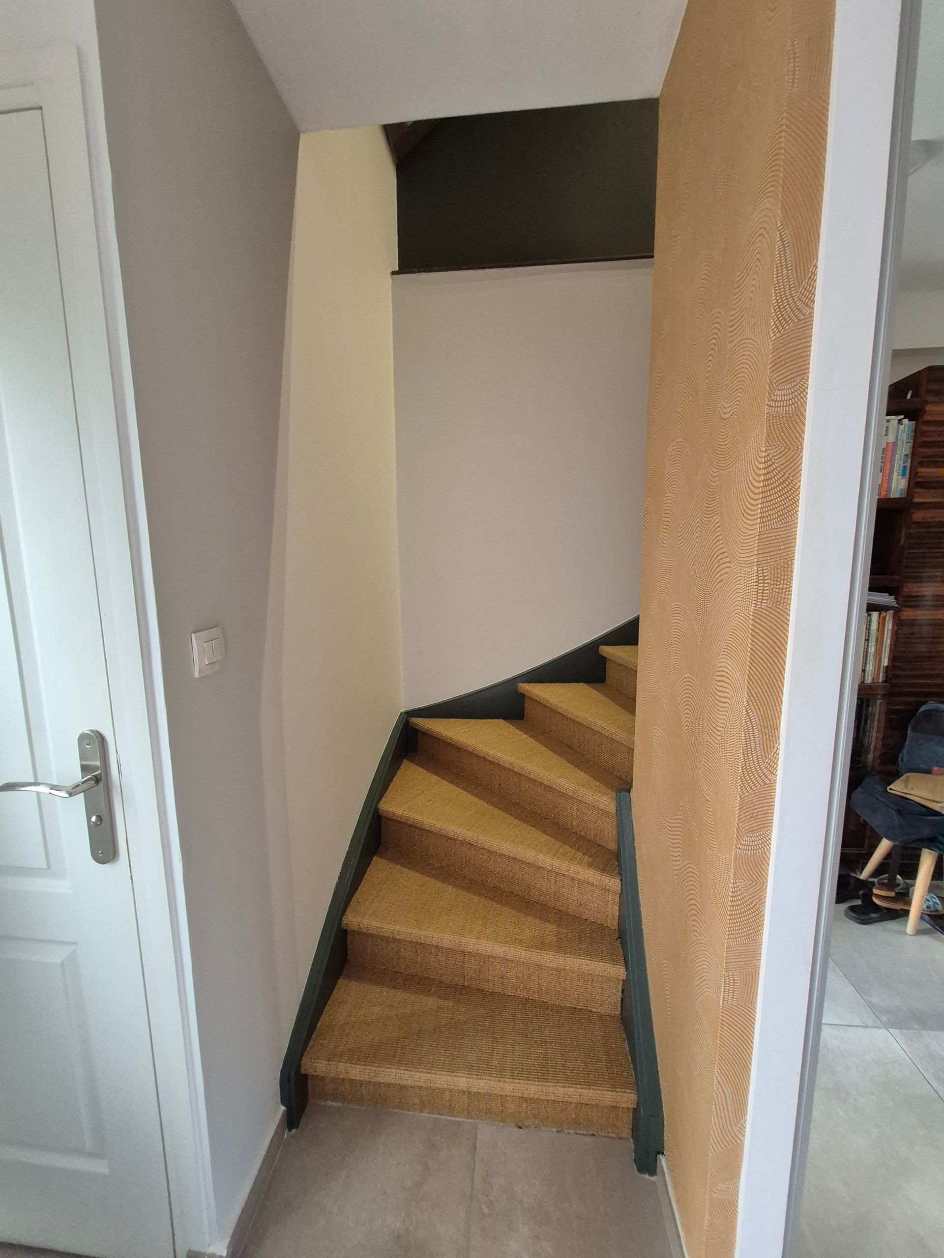 Escalier avec moquette jaune et rampe foncée, donnant sur un couloir étroit aux murs texturés.