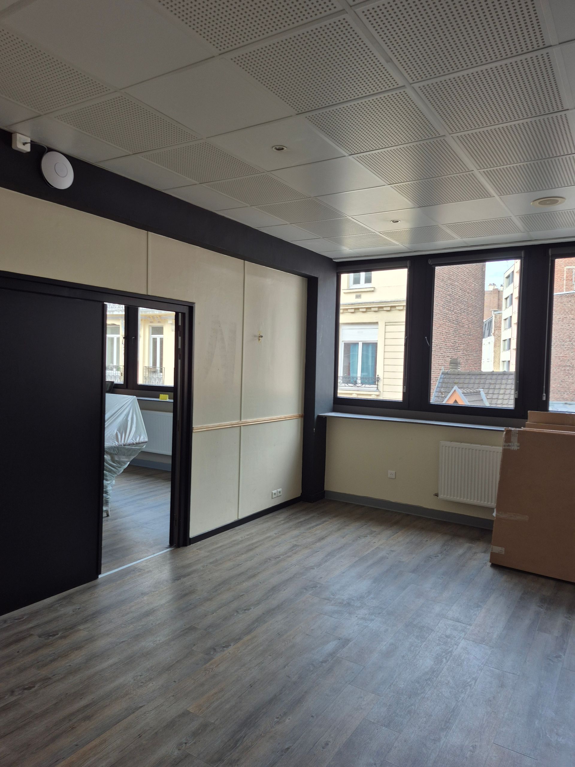 Pièce vide avec fenêtres, parquet et porte sombre. Le plafond est orné de motifs et les murs sont clairs.