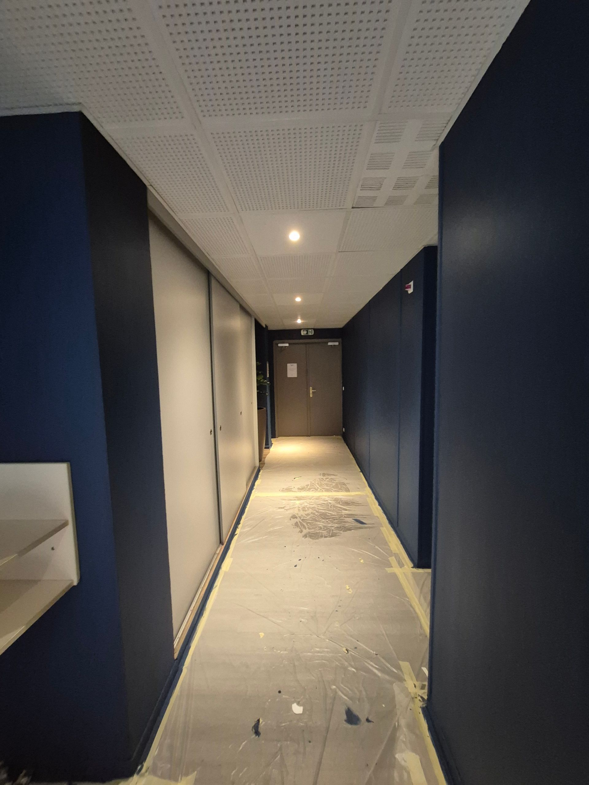 Un long couloir étroit aux murs bleus, au plafond blanc et avec une porte au fond.