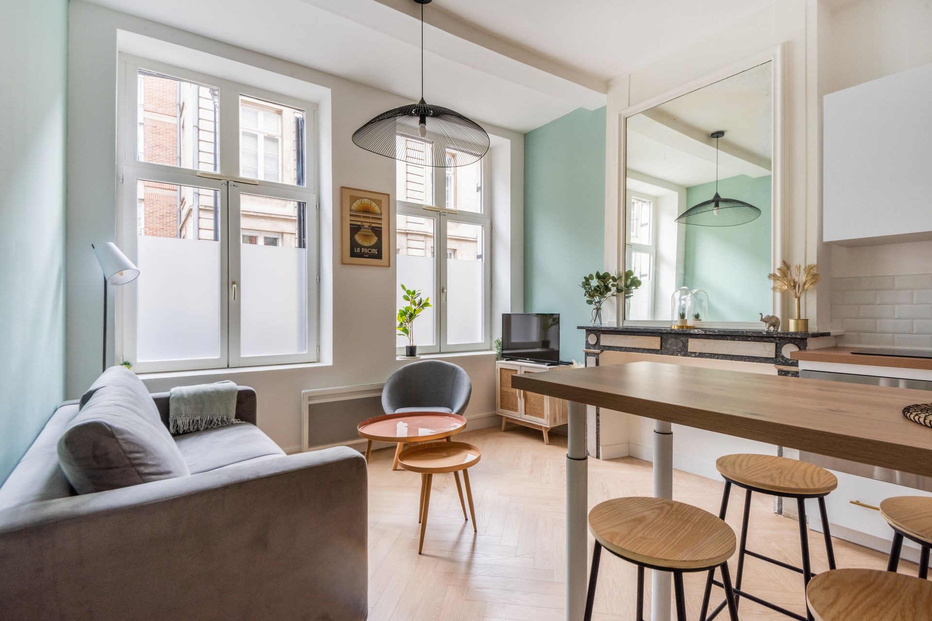 Espace de vie chaleureux avec canapé, petites tables et îlot de cuisine avec tabourets. Murs couleur menthe et grandes fenêtres.