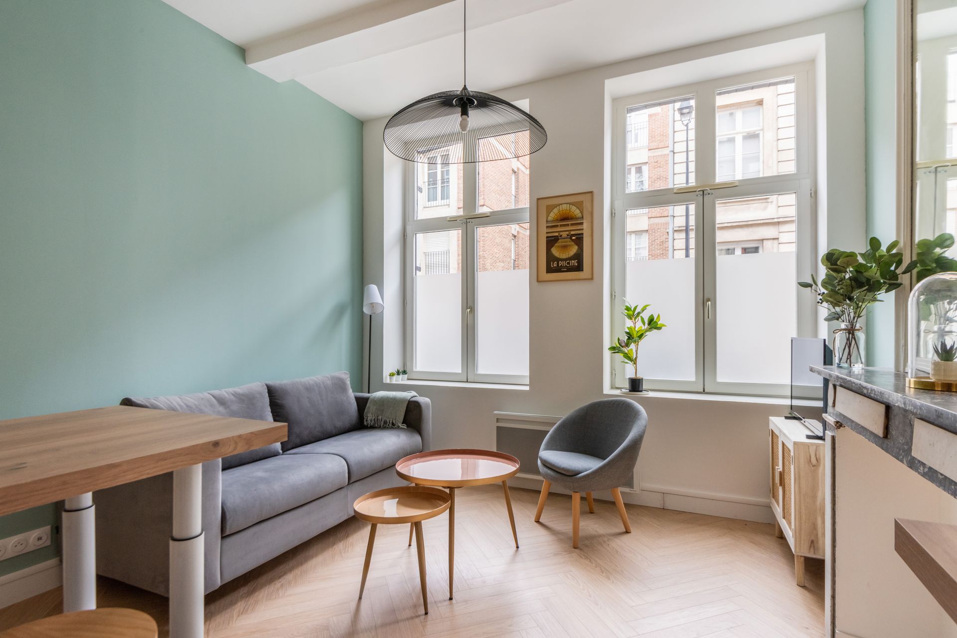 Salon avec canapé, deux fenêtres, petites tables et une table en bois. Mur bleu clair.