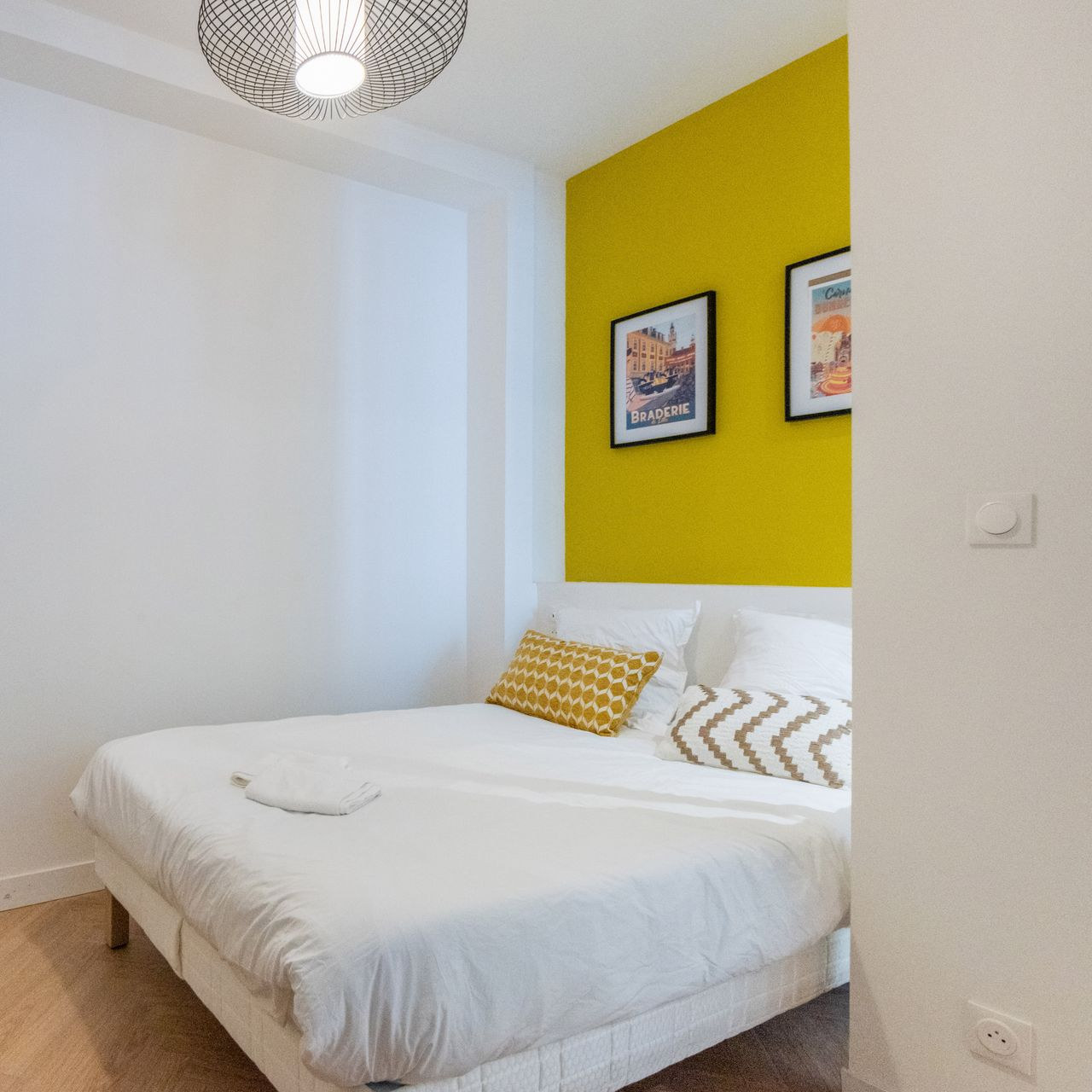 Chambre avec un mur d'accent jaune, du linge de lit blanc, des tableaux encadrés et un coussin à motifs.