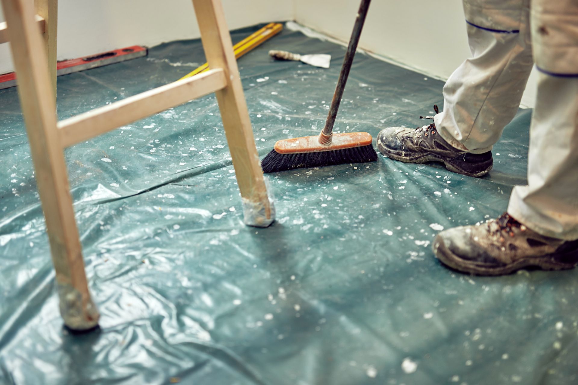 Une personne balaie des éclaboussures de peinture blanche sur un sol recouvert de plastique pendant un chantier, près d'une échelle et d'outils.