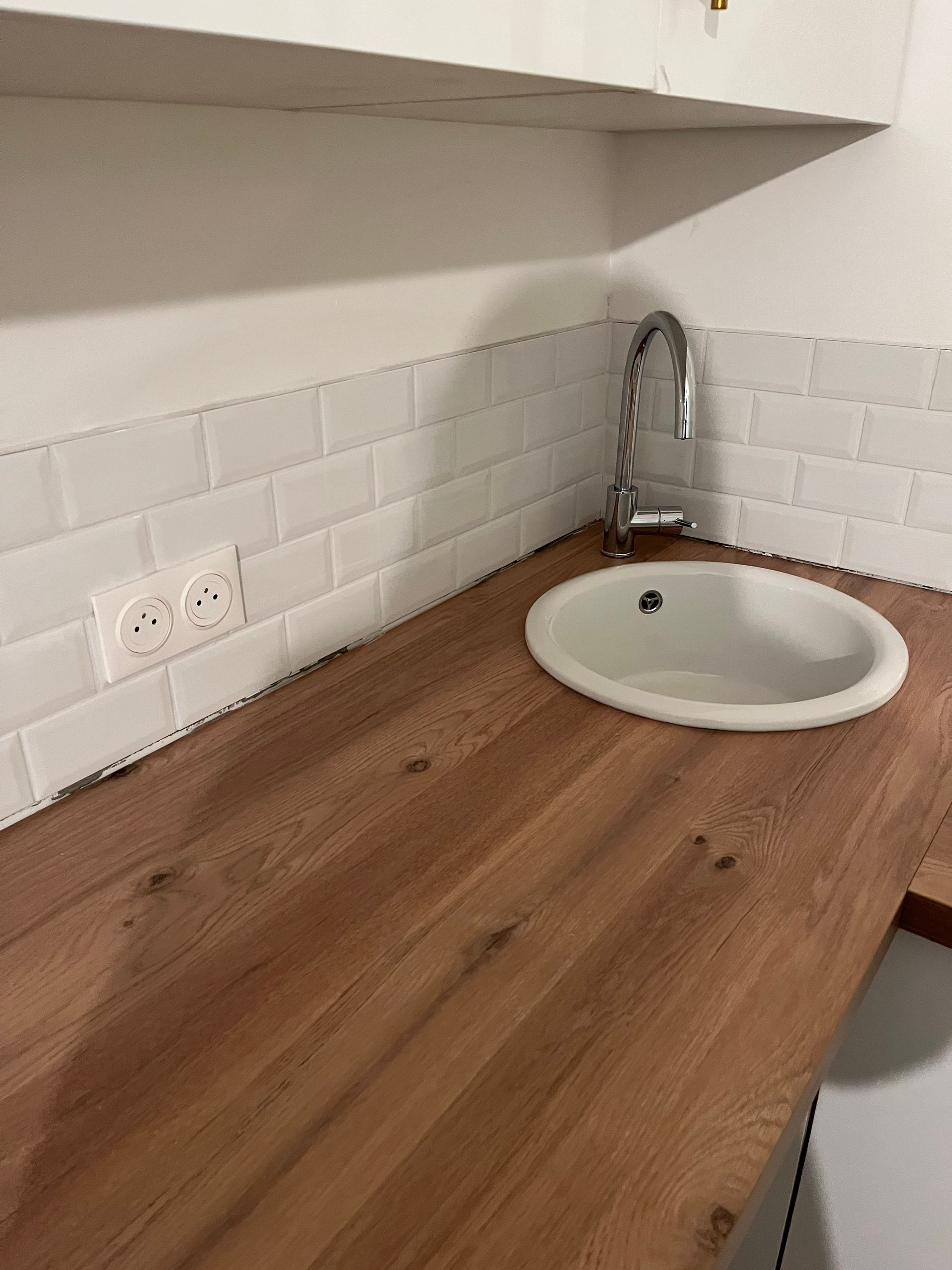 Plan de travail en bois avec évier et robinet blancs, crédence carrelée et prise électrique.