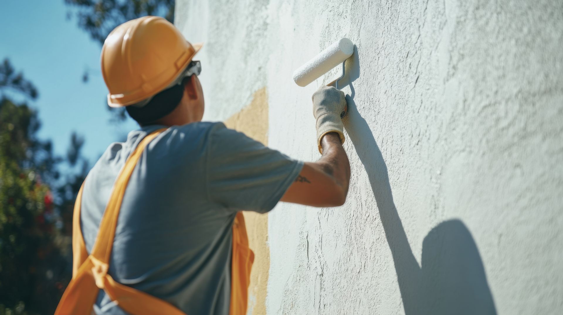 Une personne portant un casque et des gants jaunes peint un mur extérieur blanc au rouleau.