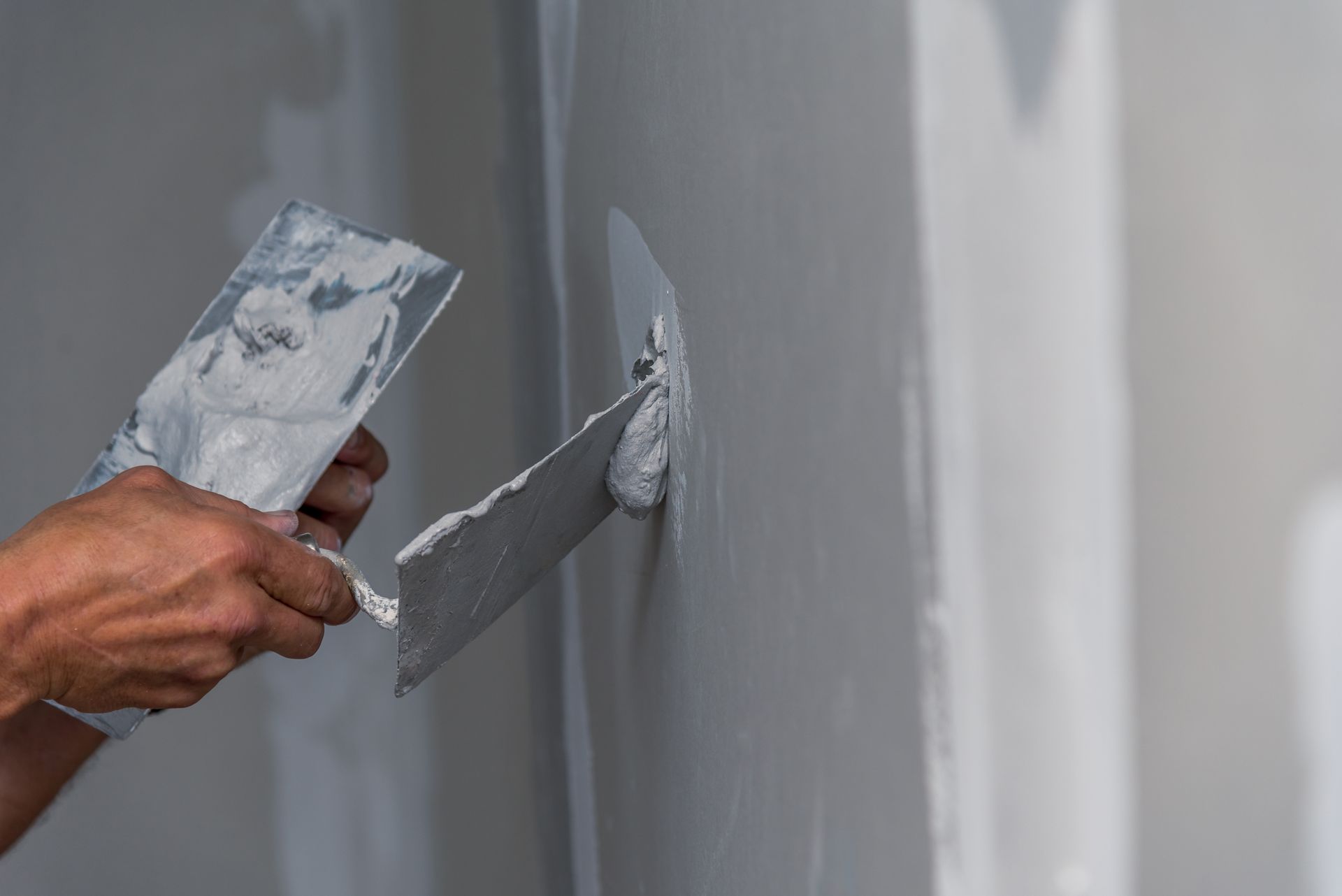 Personne utilisant une truelle pour appliquer de l'enduit à plaques de plâtre sur un mur gris clair.