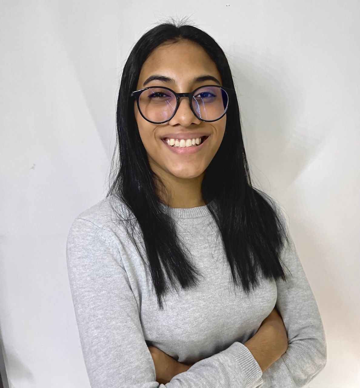 A woman wearing glasses and a grey sweater is smiling with her arms crossed.
