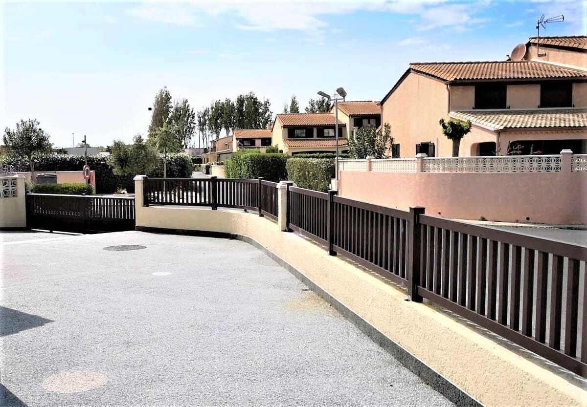 Clôture brune le long d'une allée devant des bâtiments aux couleurs claires, sous un ciel bleu.
