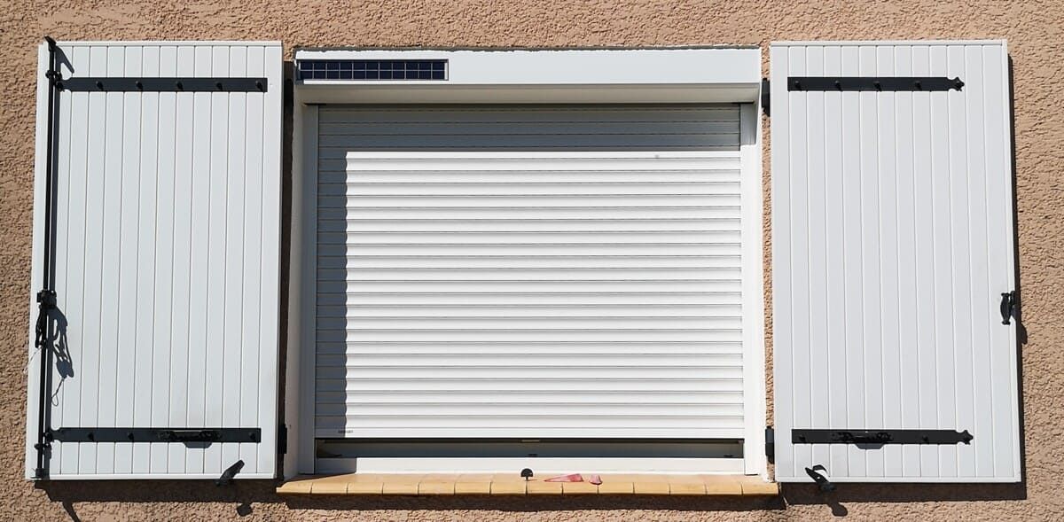 Volets blancs et stores fermés sur une fenêtre extérieure dans un mur en stuc.