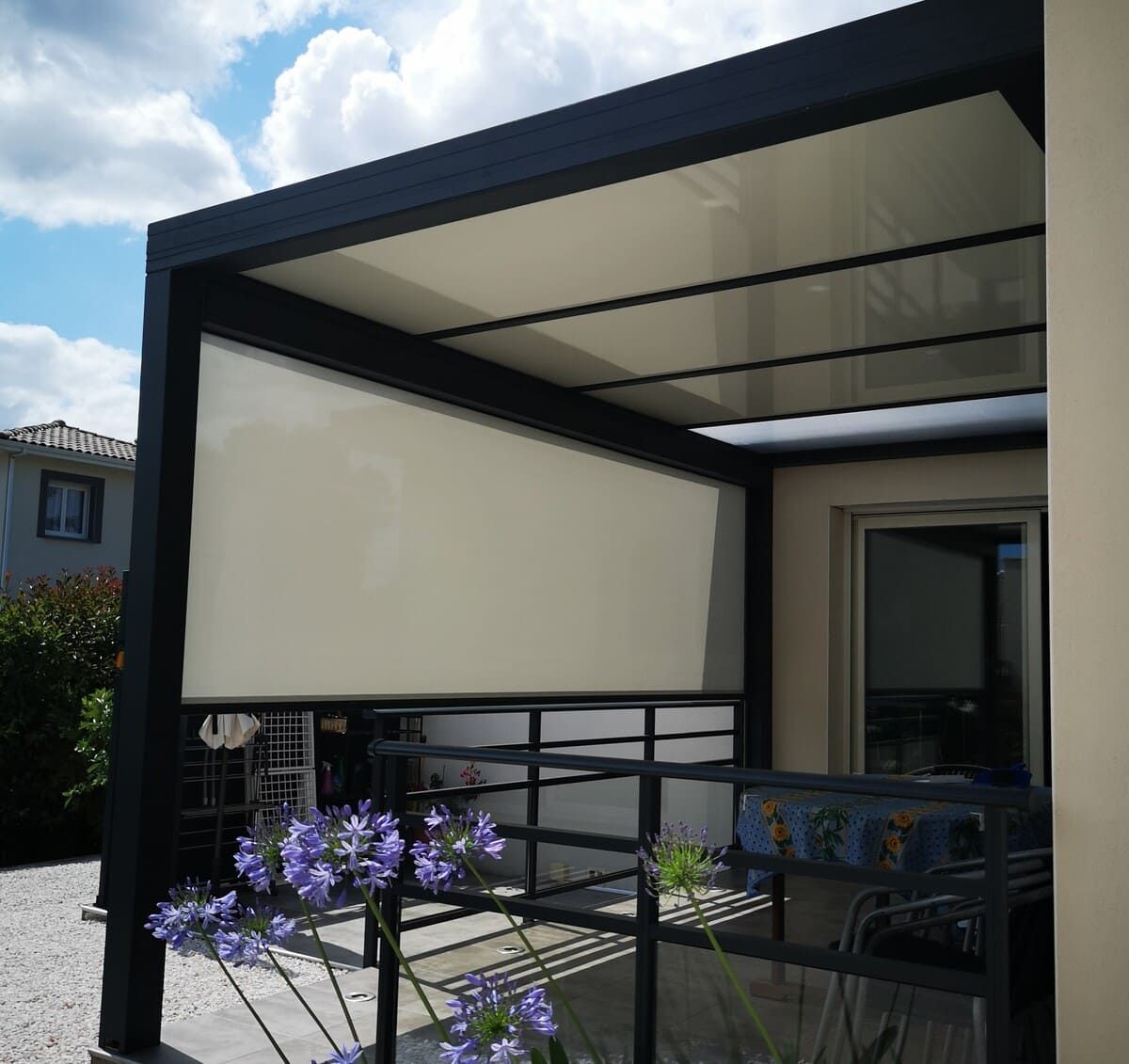 Pergola noire avec store rétractable beige, couvrant un patio orné de fleurs bleues et d'une balustrade en métal.