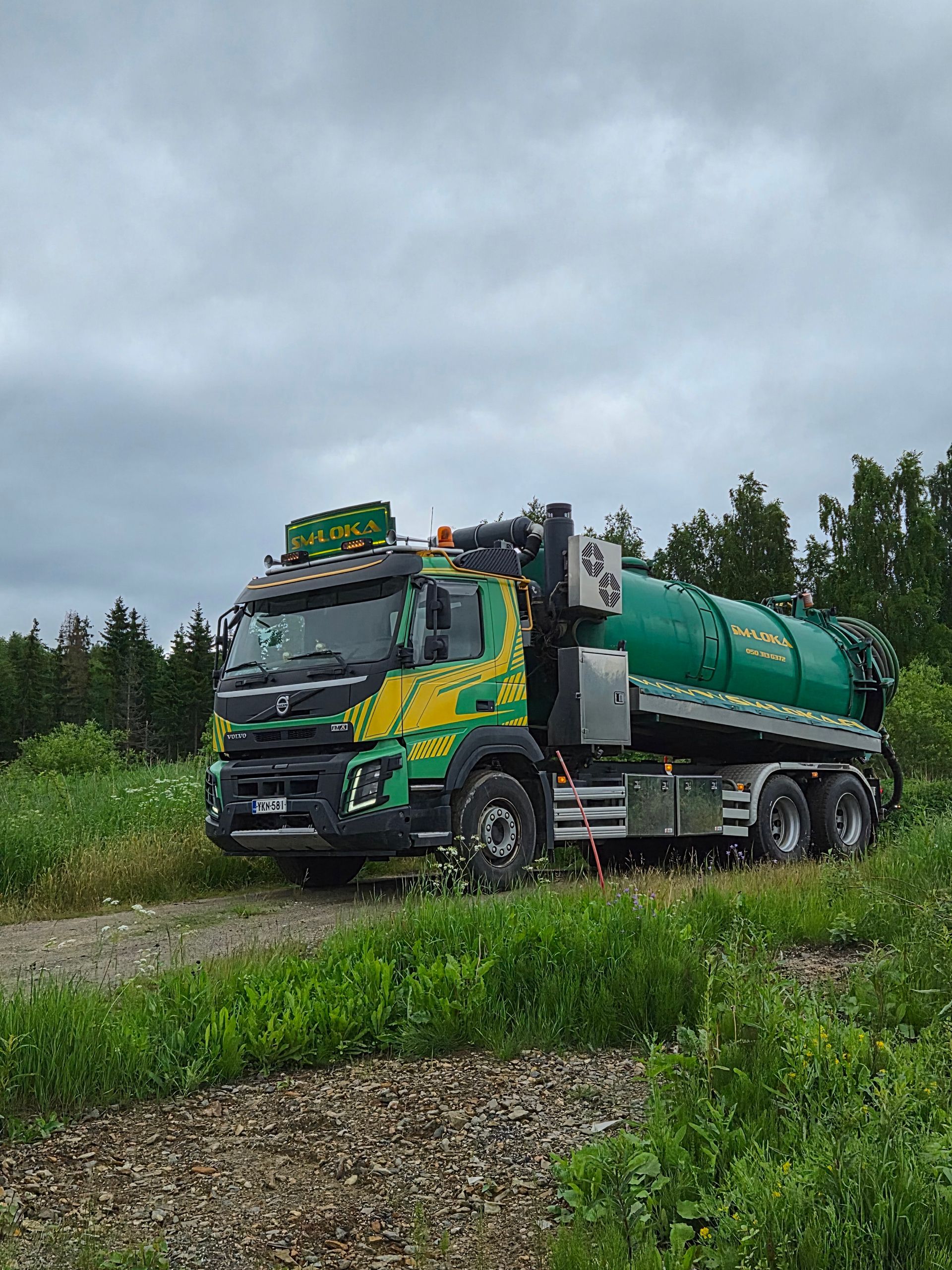 Vihreä säiliöauto sorapihalla ruiskuttamassa vettä letkulla metsäisellä pihalla.