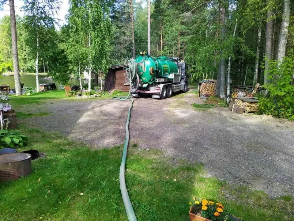 Vihreä imuriauto sorapihalla letku venytettynä metsäisen pihan poikki