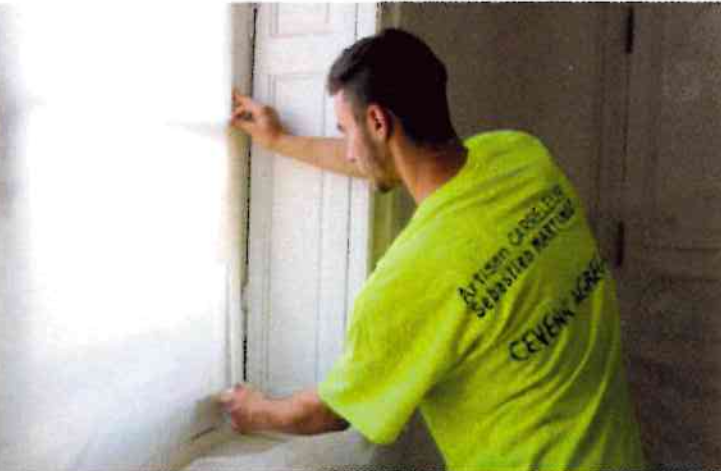 Un ouvrier vêtu d'un t-shirt vert fluo installe des moulures autour d'un cadre de fenêtre intérieur.