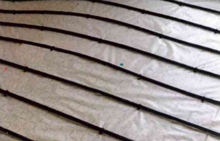 Tubes flexibles noirs pour chauffage au sol radiant, installés en rangées parallèles sur une sous-couche blanche à motif quadrillé.
