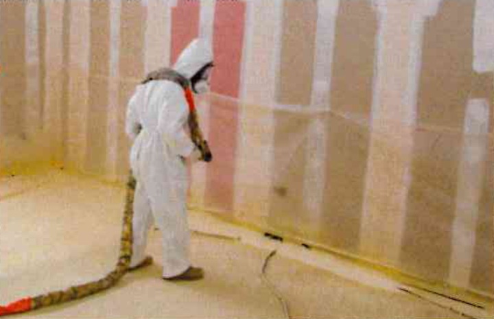 Une personne vêtue d'une combinaison de protection et d'un masque respiratoire pulvérise une substance sur un mur dans un espace intérieur.