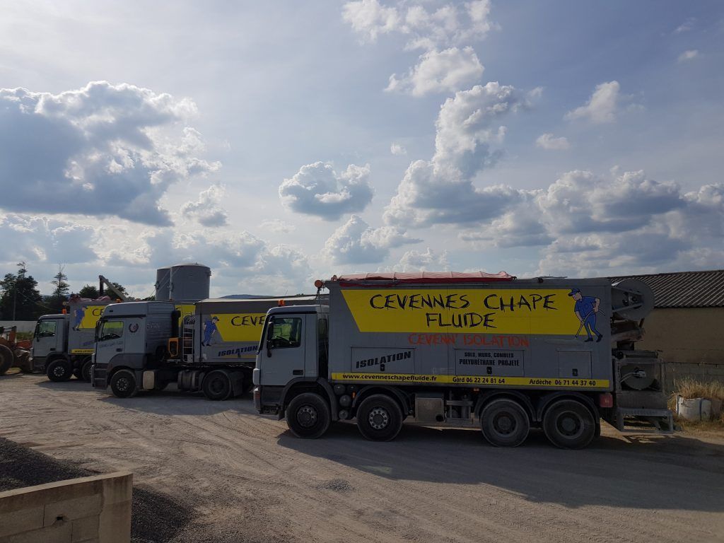 Trois camions de chantier gris sont stationnés sur un terrain gravillonné sous un ciel bleu clair et nuageux.
