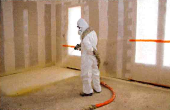 Une personne vêtue d'une combinaison de protection blanche pulvérise de l'isolant sur les murs d'une pièce inachevée tout en portant un masque respiratoire.