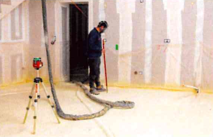 Un ouvrier utilise un outil pour lisser l'isolant sur le sol tandis qu'un niveau laser sur trépied surveille les travaux dans une pièce.