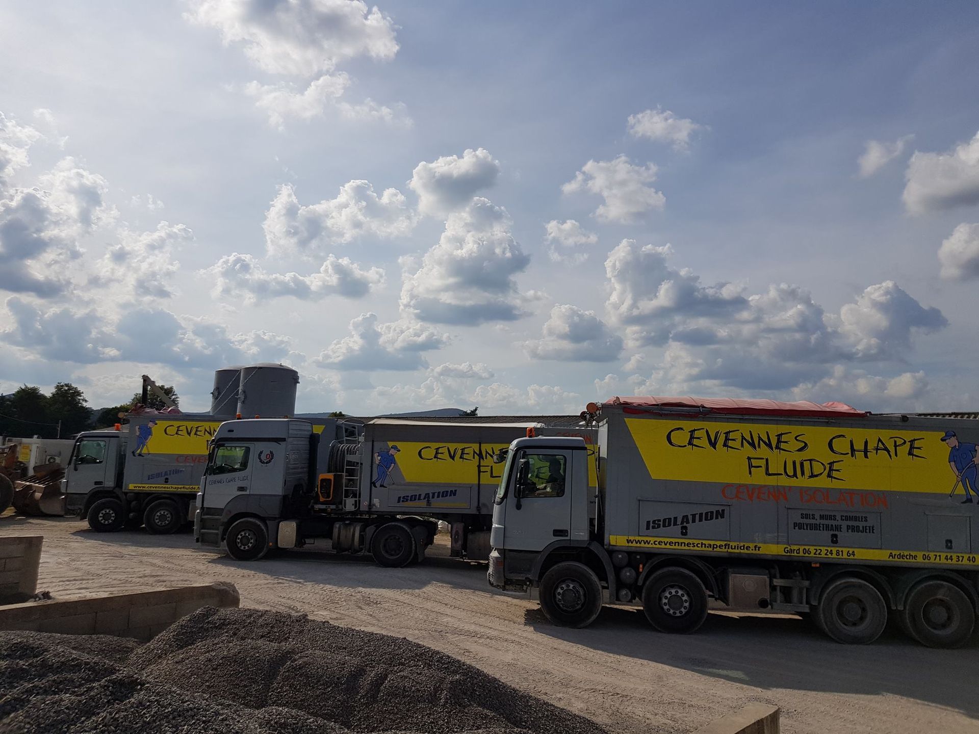 Trois camions industriels gris avec des marquages jaunes sont stationnés sur un chantier de construction en gravier sous un ciel bleu nuageux.