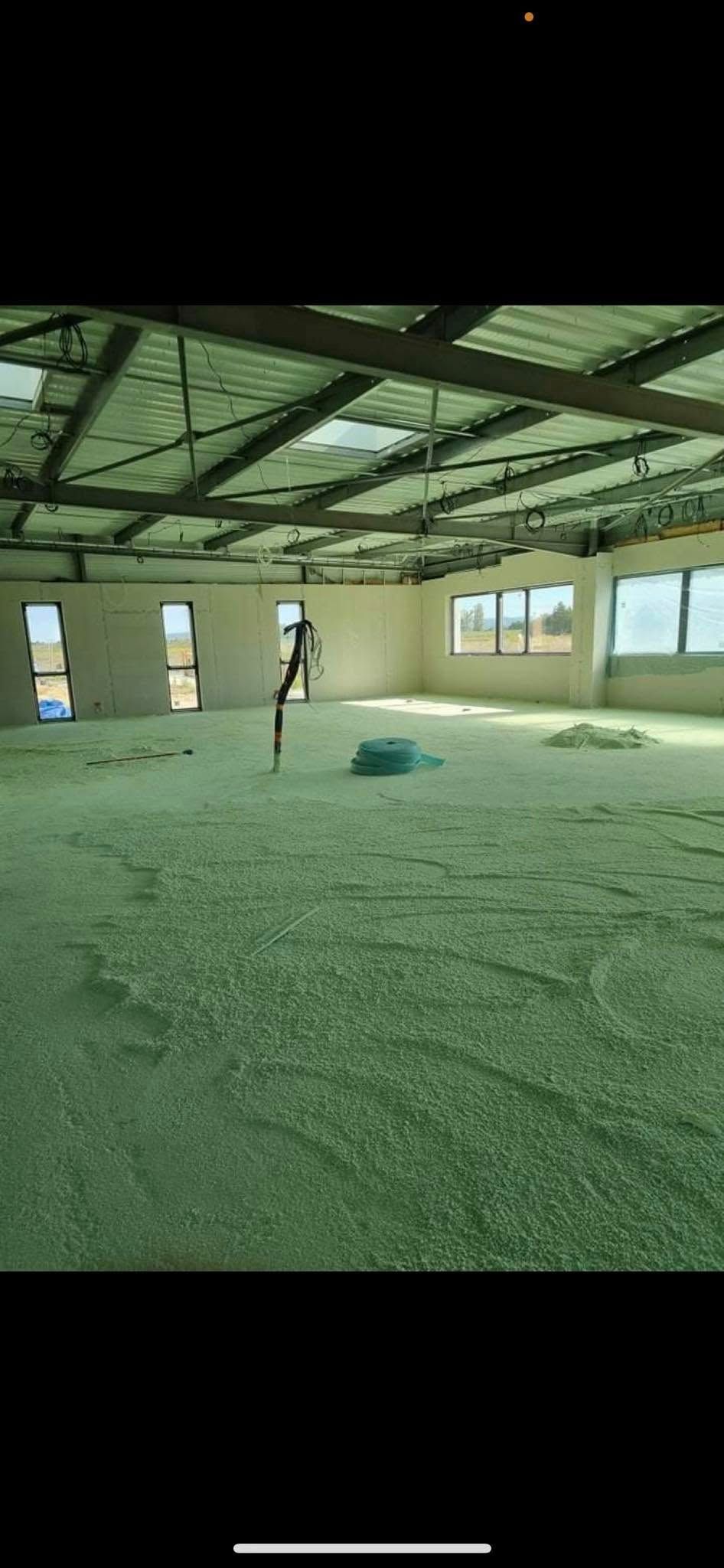 Une pièce intérieure inachevée avec un sol vert pâle, des murs blancs, de grandes fenêtres et un plafond aux poutres métalliques apparentes.