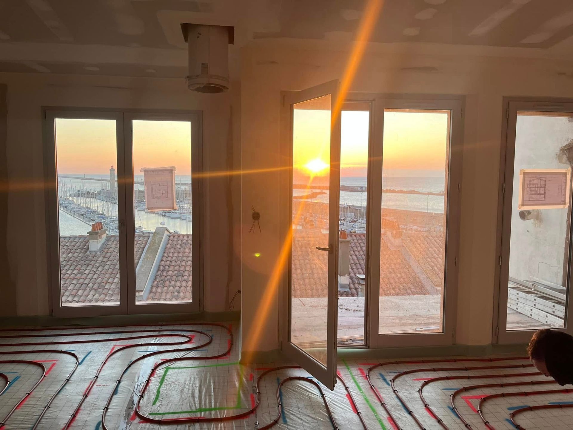 Le soleil se couche sur un port, vu depuis une pièce inachevée où l'on aperçoit des tubes de chauffage au sol.