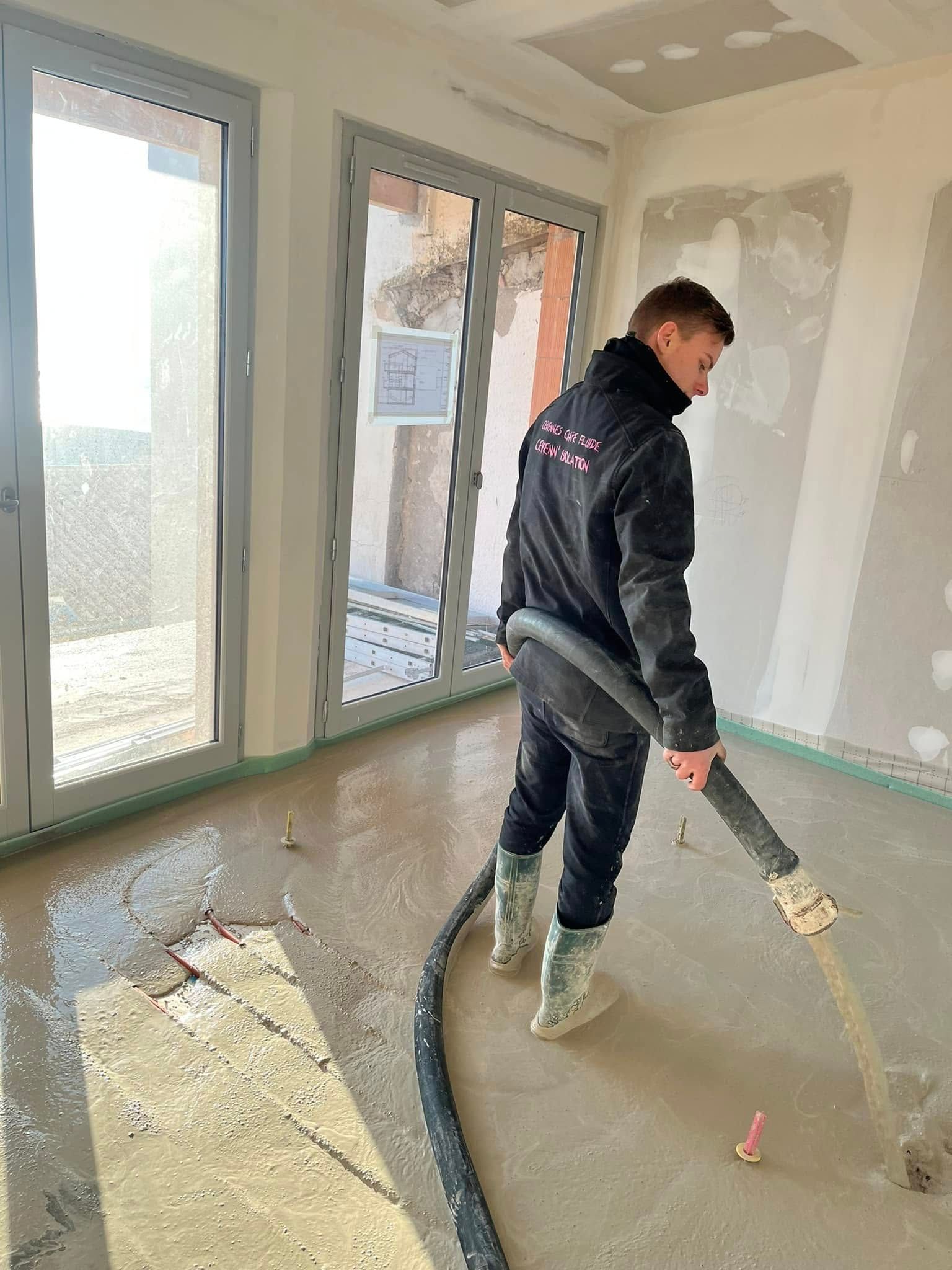 Une personne vêtue de vêtements et de bottes de travail utilise un tuyau d'arrosage pour verser du béton liquide sur le sol intérieur d'une pièce non finie.