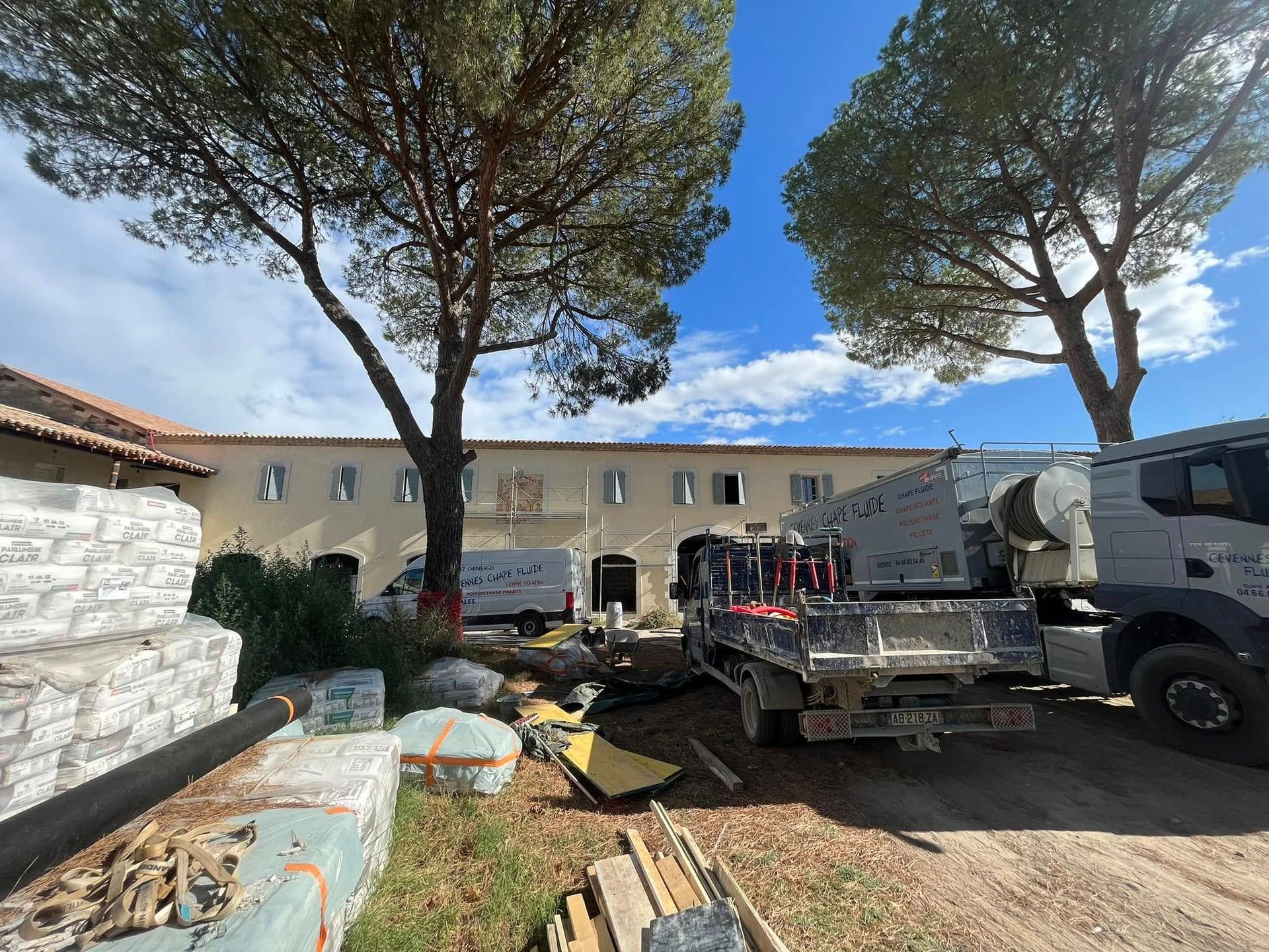 Un chantier de construction devant un long bâtiment, bordé de grands pins, sous un ciel d'un bleu limpide.