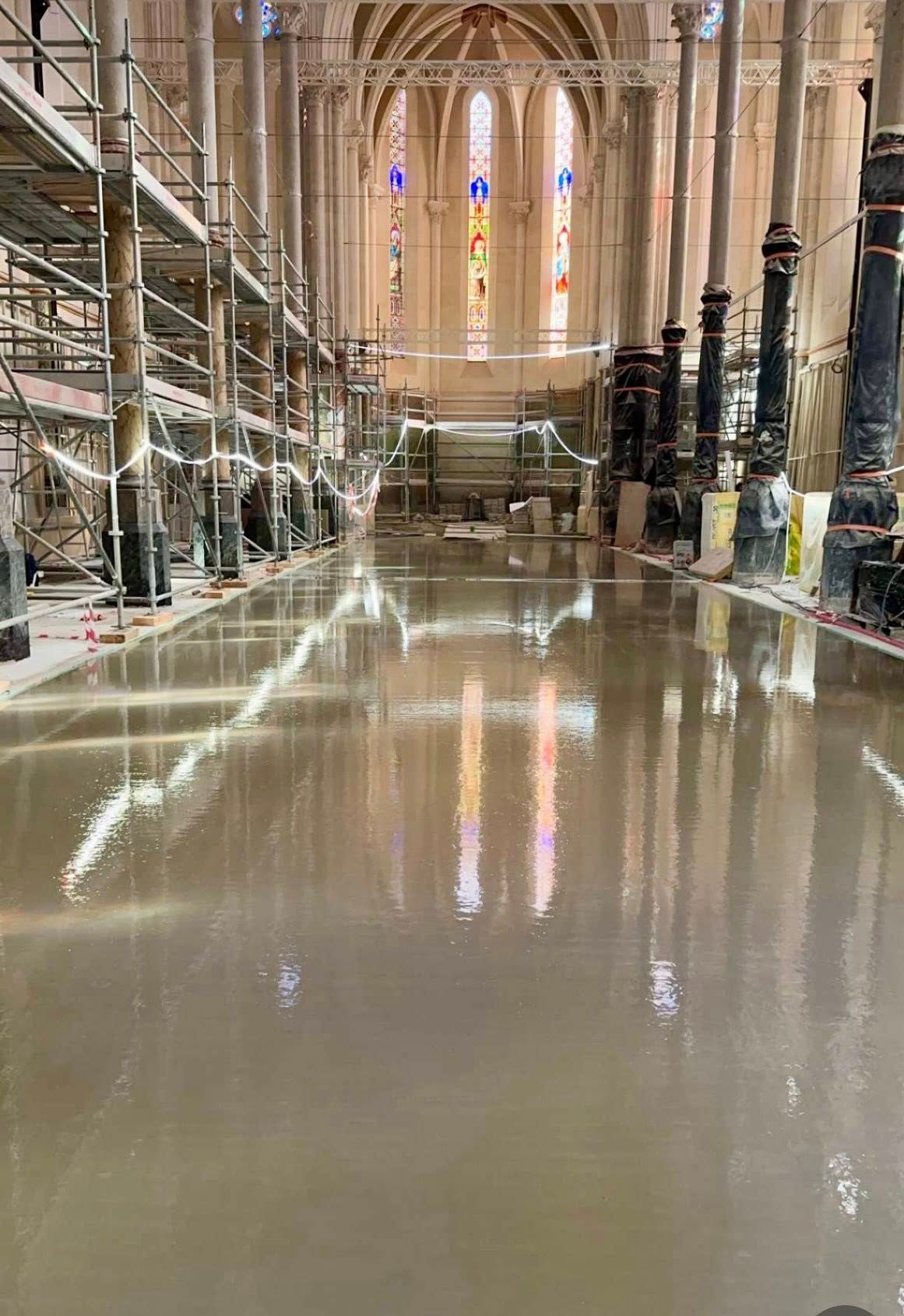 Vue en plongée de l'intérieur d'une église en rénovation, montrant des échafaudages et un sol fraîchement coulé, humide et réfléchissant.