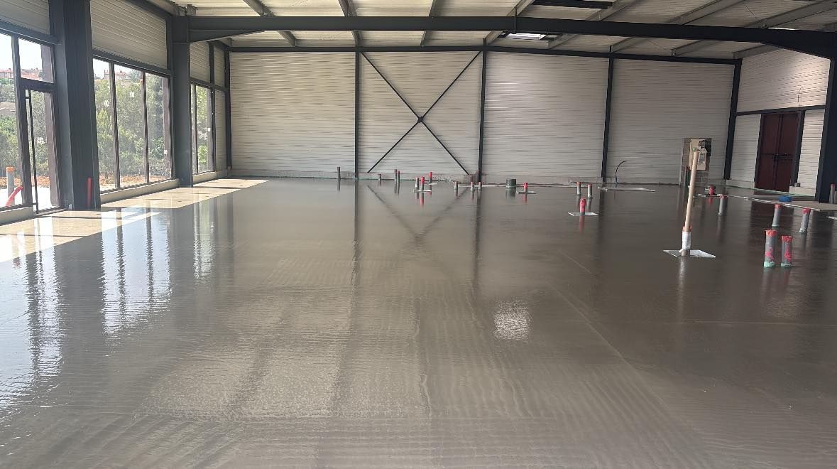 Un sol en chape fluide fraîchement coulée à l'intérieur d'un grand bâtiment à aire ouverte avec des murs en verre et des poutres de soutien en acier.
