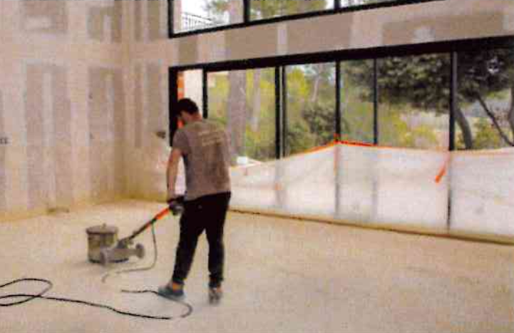 Un ouvrier utilise une ponceuse à parquet dans une pièce aux grandes baies vitrées et aux cloisons sèches non finies et jointoyées.