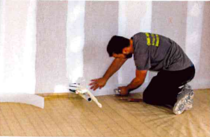 Un ouvrier s'agenouille sur une bâche pour installer des bandes isolantes le long de la base d'un mur dans une pièce.