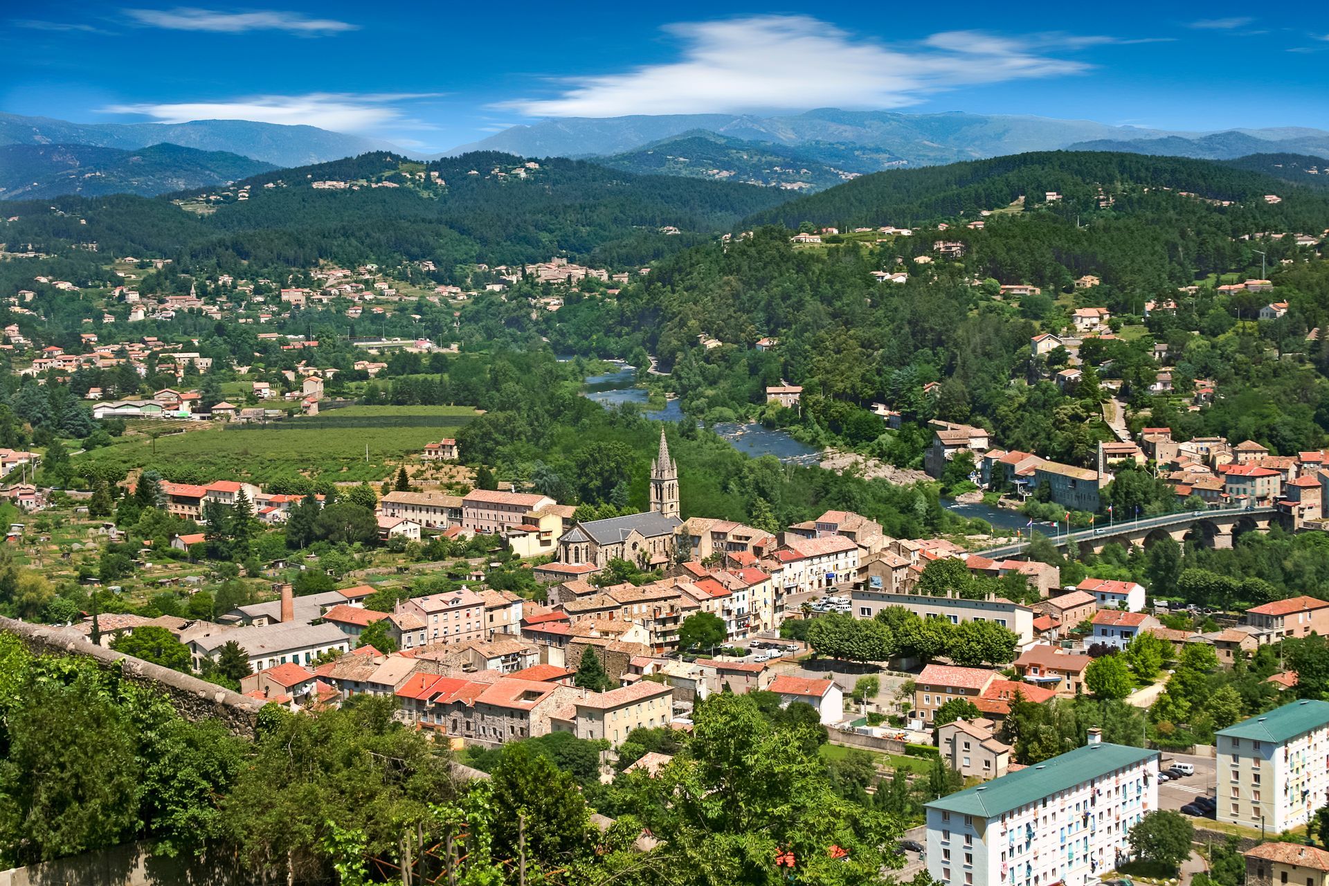 Vue en plongée d'une ville aux toits de tuiles rouges, d'un clocher d'église et d'une rivière entourée de collines et de montagnes boisées.