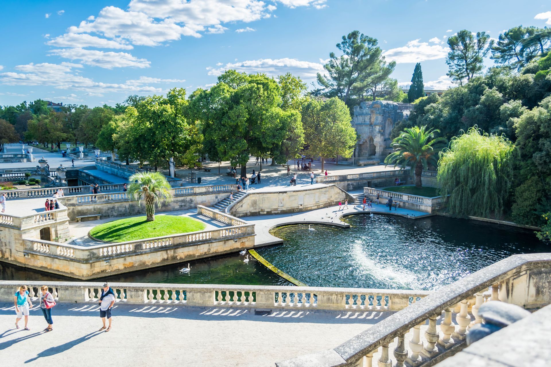 Une balustrade en pierre surplombe un parc avec un étang en contrebas, des allées aménagées, des arbres verdoyants et des promeneurs par une journée ensoleillée.