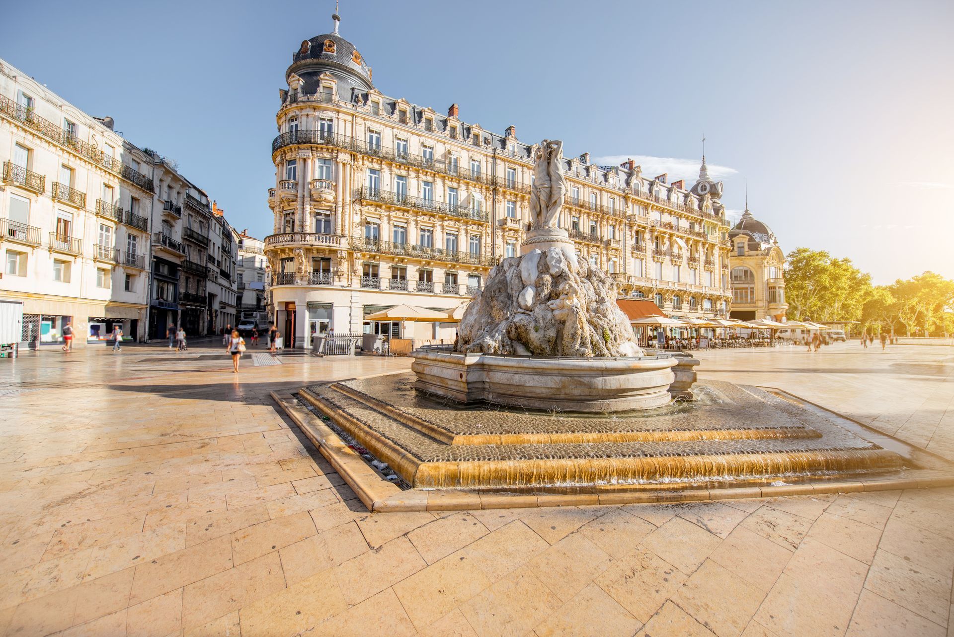 La fontaine des Trois Grâces se dresse au centre d'une place pavée et ensoleillée à Montpellier, en France, encadrée par des bâtiments historiques.