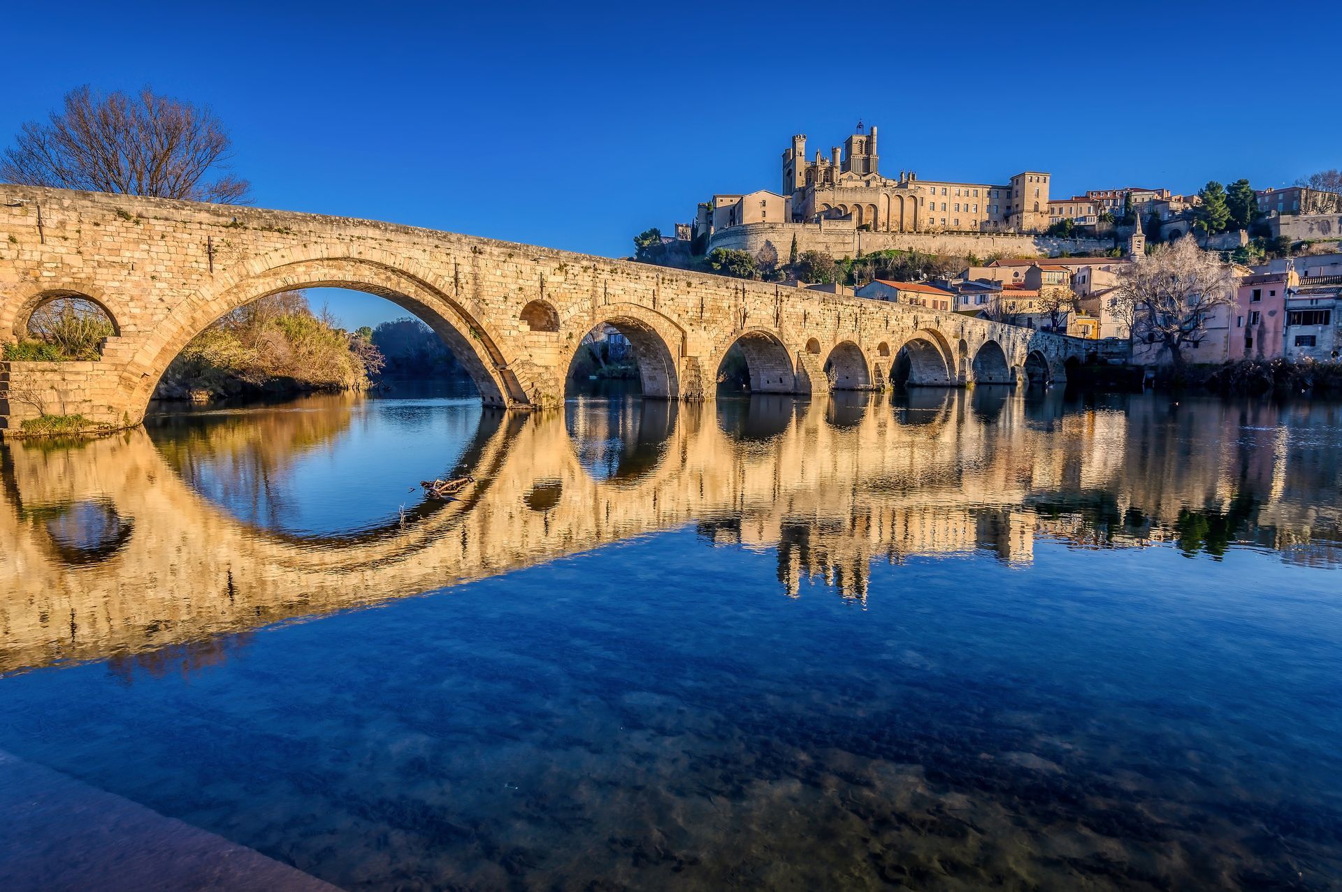 Un ancien pont de pierre à plusieurs arches enjambe une rivière calme et miroitante sous un ciel d'un bleu limpide, au-dessus d'une ville historique.