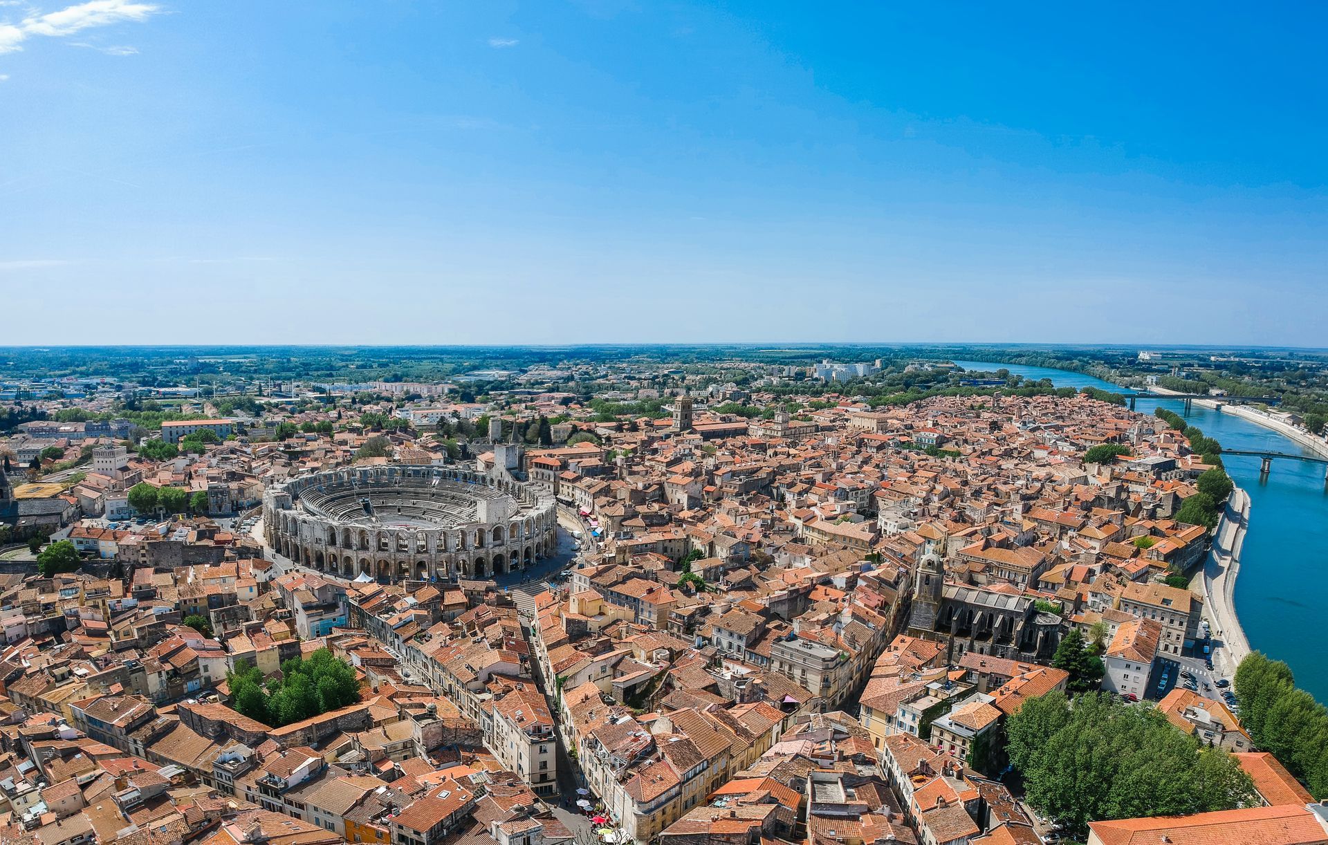 Vue aérienne des arènes romaines d'Arles, en France, entourées d'une ville dense aux bâtiments aux toits orangés, au bord d'une rivière.