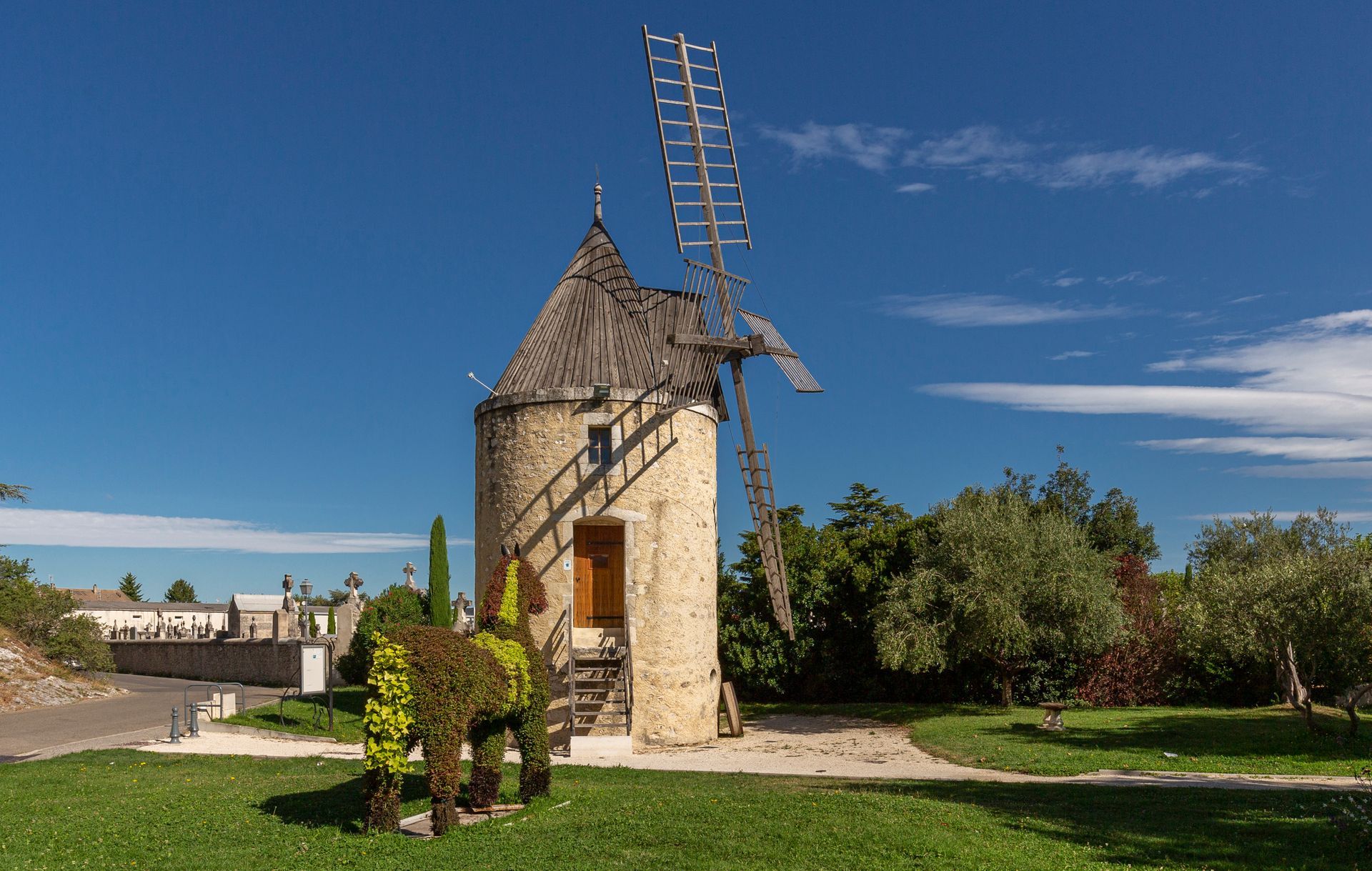 Un moulin à vent en pierre, au toit en bois et aux pales partiellement manquantes, se dresse sur une pelouse herbeuse à côté d'un animal taillé en topiaire.