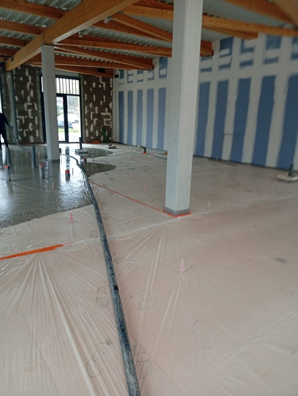 Un espace intérieur inachevé avec un sol en béton, des piliers de soutien blancs et des murs recouverts de panneaux de plaques de plâtre bleues enduites.