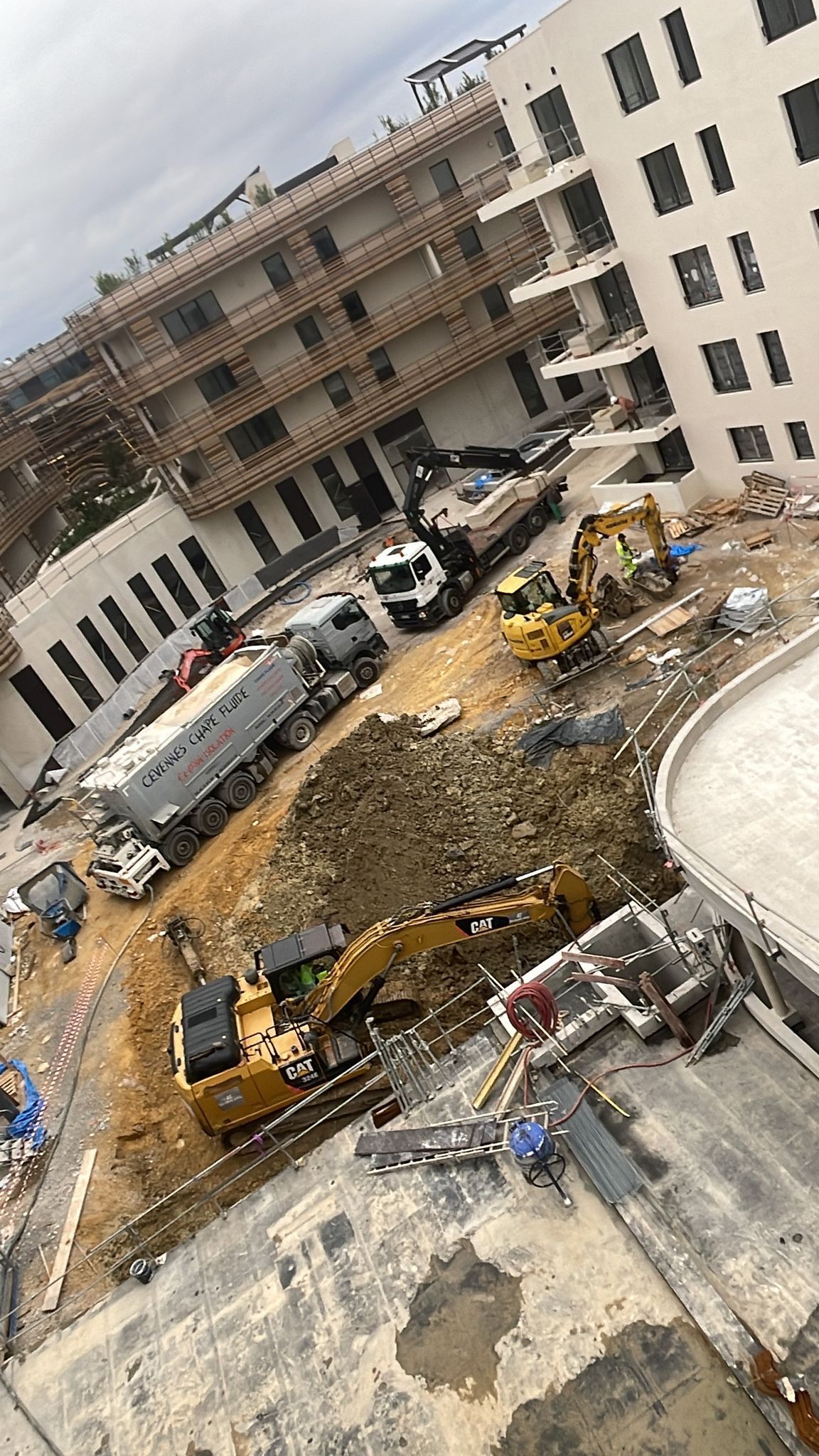 Chantier de construction avec des excavatrices jaunes et un camion malaxeur à béton travaillant sur un terrain vague entre deux bâtiments.