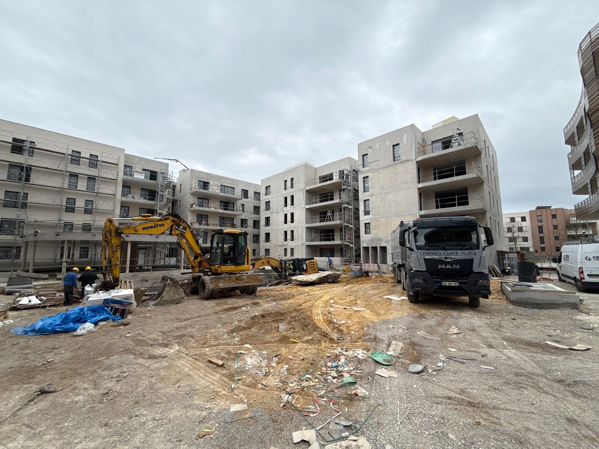 Chantier de construction avec une pelleteuse jaune et un camion gris garés devant des immeubles résidentiels modernes en cours de construction.