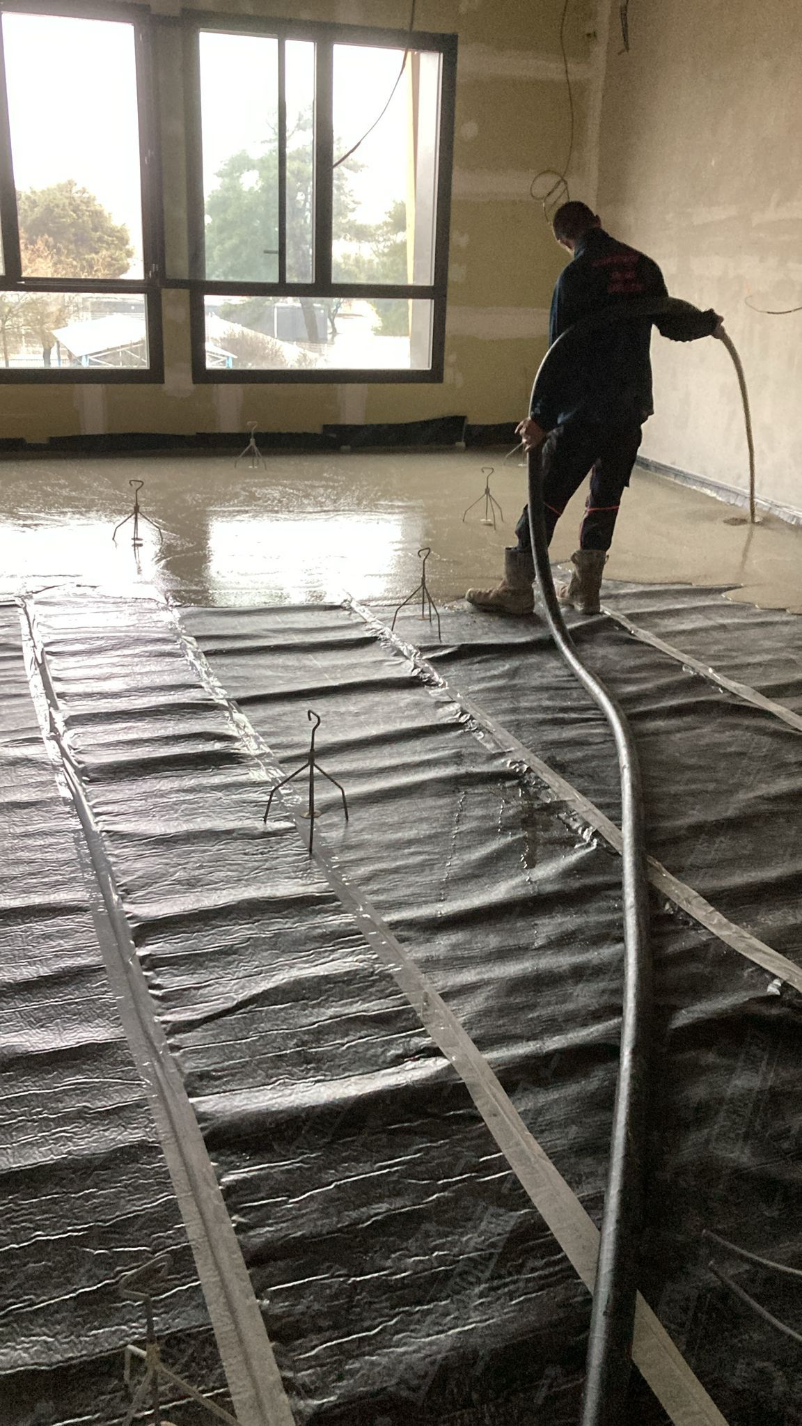 Un ouvrier pulvérise un enduit de nivellement sur un sol recouvert de tapis chauffants radiants dans une pièce dotée de grandes fenêtres.