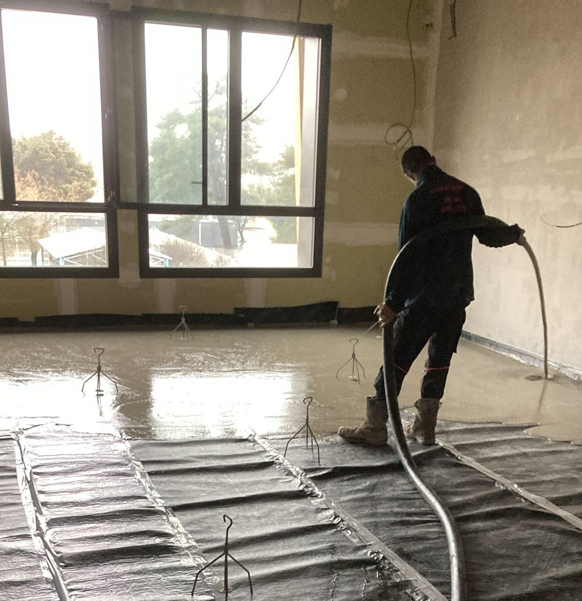 Un ouvrier utilise un tuyau pour verser un enduit de sol autonivelant sur une sous-couche sombre à l'intérieur d'une pièce dotée de grandes fenêtres.