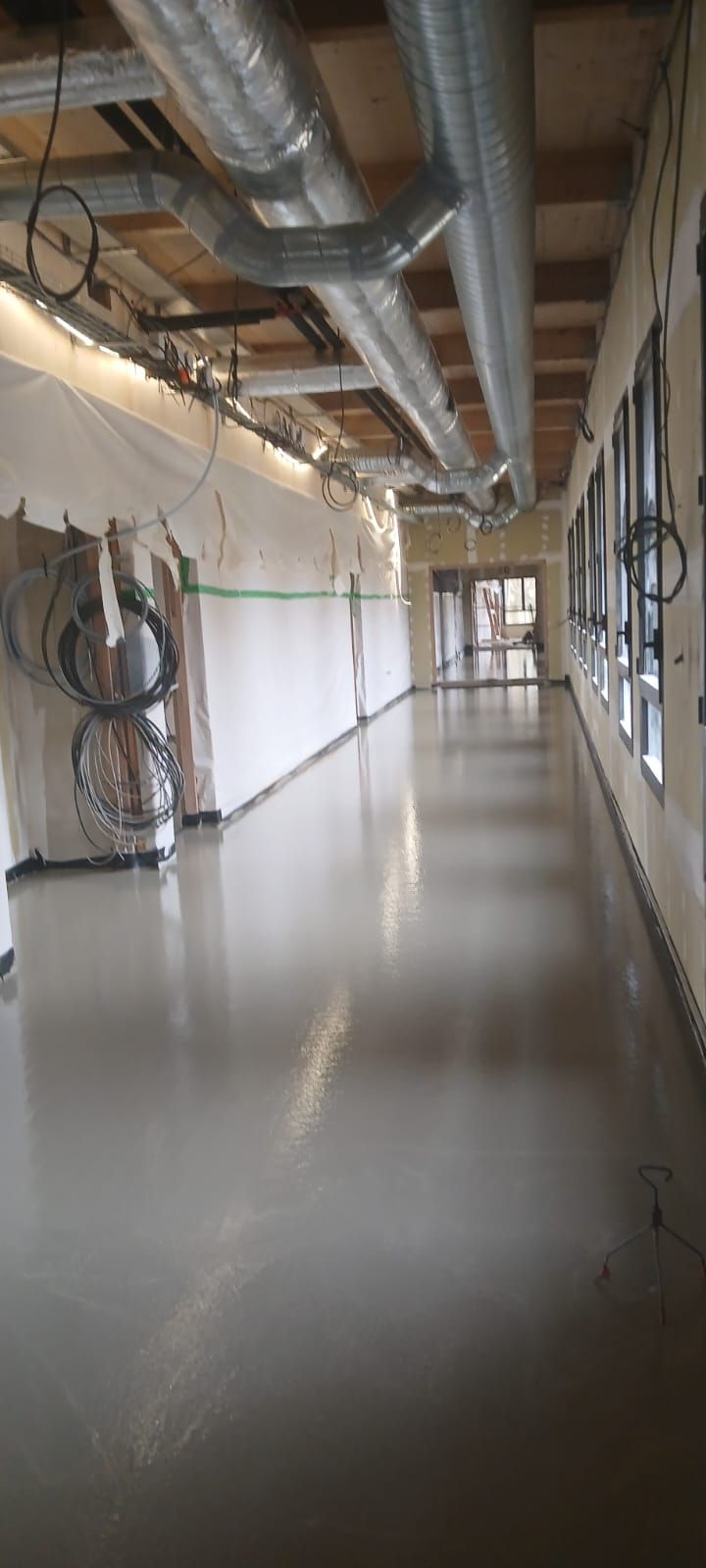 Un long couloir en construction avec un sol gris clair réfléchissant, des conduits de ventilation apparents au plafond et des parois recouvertes de plastique.
